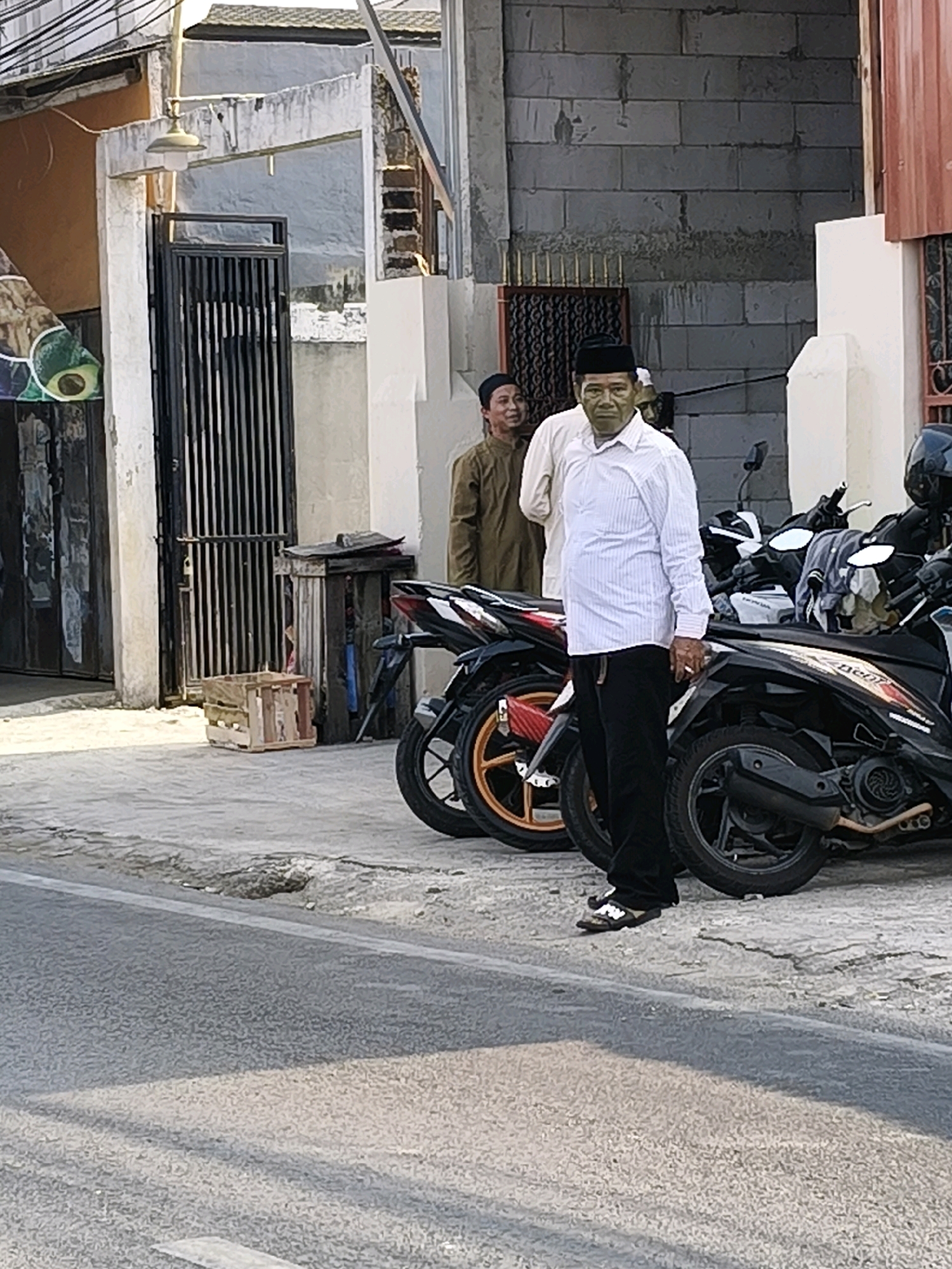 The image depicts an outdoor scene in Karangsatria, Indonesia. In the foreground, a man in a white pinstriped long-sleeved shirt and black pants stands on a cracked asphalt surface next to several motorcycles. He wears a black cap and sandals. Behind him, another man in a brown garment and a black cap is visible. The background includes a building with a gray brick wall and a white facade, a black metal gate, and a wooden crate. The lighting suggests it is daytime.