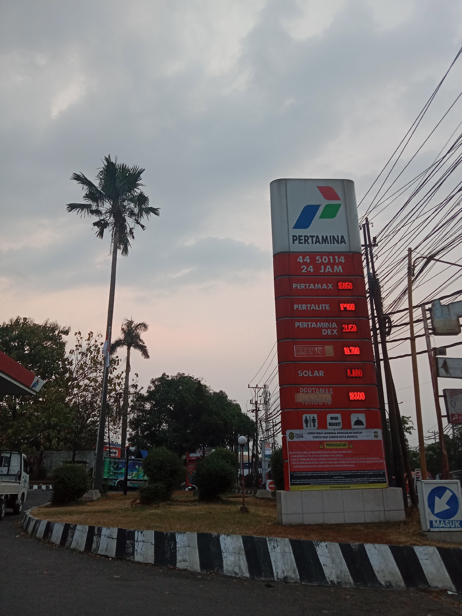 The image shows a Pertamina gas station sign with fuel prices displayed. The sign is tall and red, with the Pertamina logo at the top. Below the logo, the sign lists prices for Pertamax, Pertalite, Pertamina Dex, and Solar. There is also a phone number and "24 JAM" (24 hours) displayed. The sky is overcast. In the foreground, there is a road with black and white striped borders. In the background, there are trees, a truck, and power lines.
Based on the presence of Pertamina, a state-owned oil and gas company in Indonesia, and the Indonesian language on the sign, this scene is likely located in Indonesia. While the exact location is not explicitly stated, Pertamina gas stations are common throughout Indonesia, including in cities like Semarang. The image captures a typical street view of a gas station in an urban or suburban area. There are no people clearly visible in the image.
