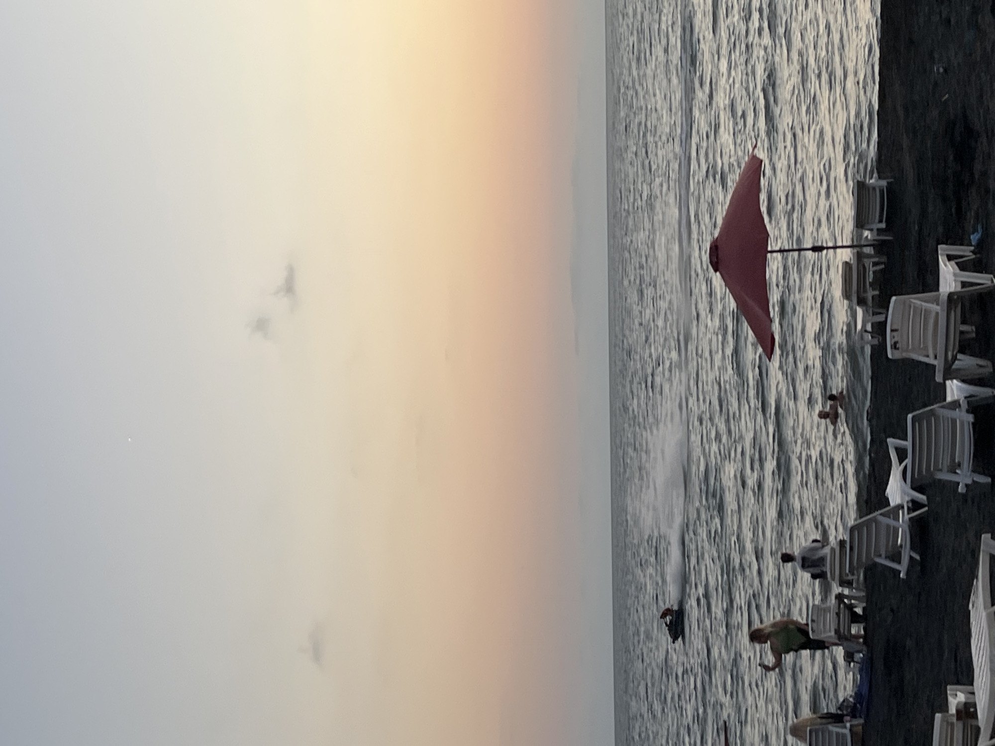 A panoramic view of the Black Sea coastline at dusk in Ureki, Georgia. The sky transitions from a soft pink to a pale yellow, with faint clouds scattered across the horizon. The sea is choppy, with small waves reflecting the dimming light. In the mid-ground, a person is riding a jet ski, leaving a trail of white spray. Several people are visible near the water's edge. Some are wading or swimming, while others are standing on the dark sand beach. In the foreground, a bright red umbrella shades two empty lounge chairs. More white lounge chairs are scattered across the beach, some occupied and some empty. The scene captures a peaceful evening at a popular coastal resort.