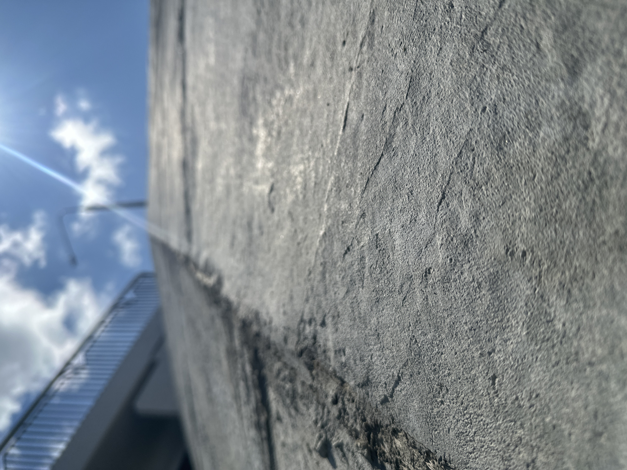 This is a close-up, low-angle shot of a textured concrete surface, likely a ramp or elevated walkway, under a bright, sunny sky with scattered clouds. The sun creates a lens flare in the upper right quadrant of the image. In the background, a metal railing and a portion of a light-colored structure are visible, out of focus. The concrete shows signs of wear, with darker, rougher patches and linear indentations. The image does not depict any people or specific landmarks that would allow for identification of a location within Minsk, Belarus.