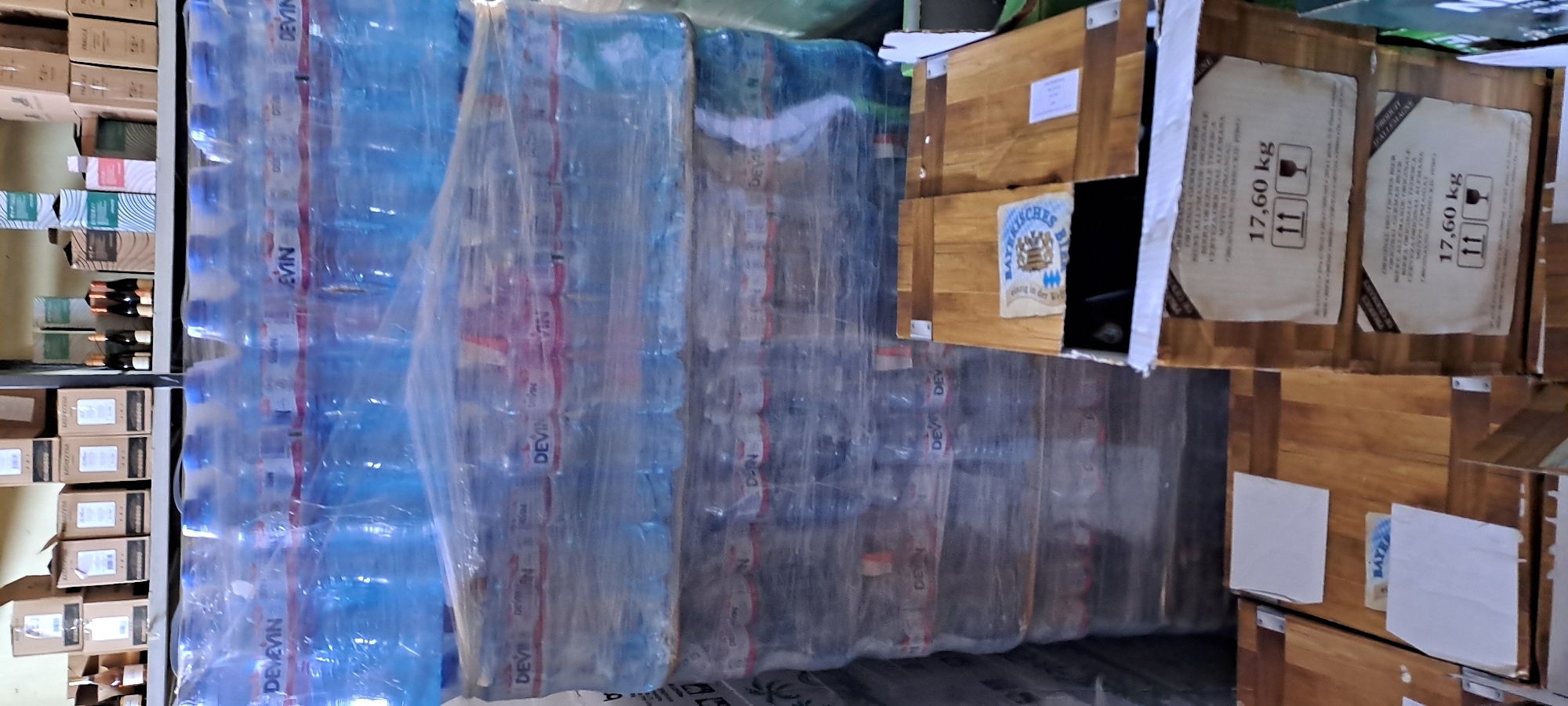 The image depicts a storage area in Velika, Bulgaria, likely a warehouse or a shop's backroom. The foreground is dominated by stacked cases of bottled water, shrink-wrapped on pallets, with the brand "DEVIN" visible on the labels. To the right, there are wooden crates, some with labels indicating "17,60 kg" and "ORIGINAL GERMAN BEER". In the background, shelves hold various boxes and bottles, including what appear to be wine bottles and other packaged goods. The lighting is somewhat dim, suggesting an indoor environment with artificial or limited natural light. There are no people visible in the frame.