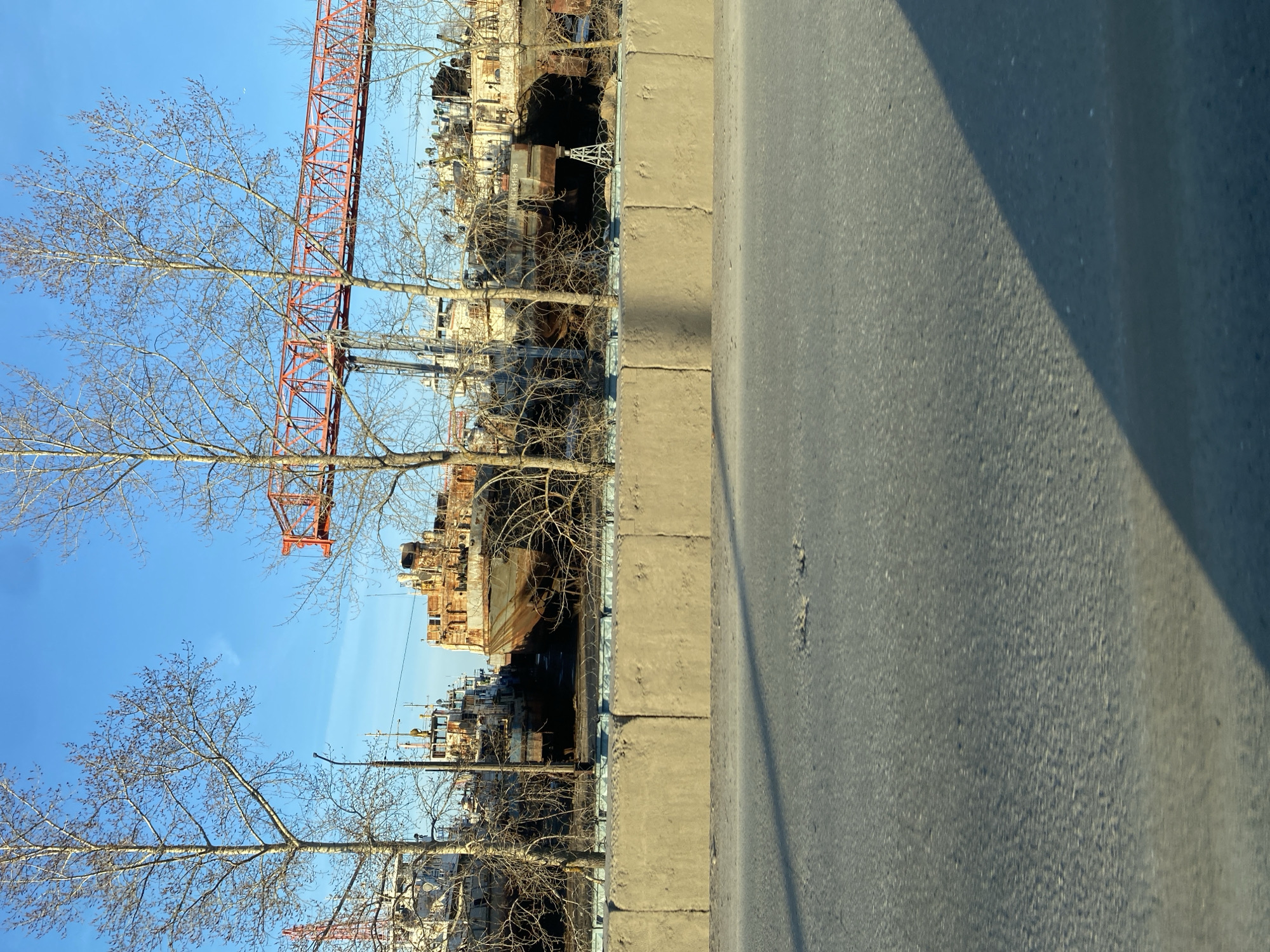 This media file captures a waterside industrial scene in Chaikovsky, Russia, likely during late autumn, winter, or early spring. The photograph is taken from what appears to be a road or pavement, which occupies the right side of the frame. This asphalt surface shows patterns of light and shadow, suggesting bright sunlight. A sturdy concrete barrier, possibly a curb or retaining wall, runs horizontally across the middle ground, separating the road from a lower area.
Beyond this barrier, a line of deciduous trees with bare branches stands, their leafless state emphasizing the skeletal structure against a clear, bright blue sky. Through and above these trees, a series of large, rusty, industrial vessels, possibly old ships or barges, are visible, hinting at a shipyard, port, or shipbreaking facility. The ships appear weathered and heavily corroded, with visible superstructures and hulls. One vessel prominently displays the Cyrillic text "ПК-58" on its side.
Dominating the upper left portion of the image, stretching across the sky and partially obscured by tree branches, is a massive red metal structure, identifiable as a lattice-boom crane or a section of an industrial gantry. Its intricate framework stands out against the clear sky, reinforcing the industrial character of the setting. The overall impression is one of a dormant or working industrial landscape under bright, crisp daytime conditions, devoid of visible human activity.