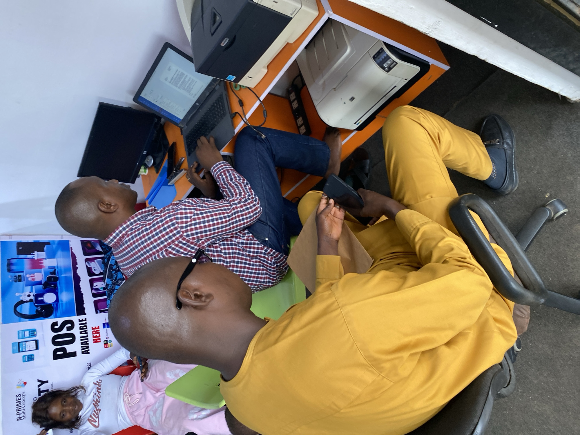 This image depicts an indoor scene at a multi-service business center or office in Bukuru, Nigeria, featuring three individuals and various electronic devices. The perspective is from slightly above, looking down into the room.
In the foreground, a man dressed in a yellow long-sleeved shirt and matching trousers is seated on a black office chair, holding a black smartphone in his hands. He wears black loafers and has black-rimmed glasses positioned above his ears. Next to him, another man, wearing a red and white checkered shirt and blue jeans, is actively using a black laptop placed on an orange-colored desk. An external monitor connected to the laptop displays text, and a black desktop computer tower sits on a shelf above. Below the desk, a power strip and cables are visible.
To the right, two beige or white printers are positioned on orange-colored shelves. The flooring is dark, possibly concrete.
On the left side of the image, a young girl lies on a pink blanket or clothing on the floor. She wears a white long-sleeved shirt with "NuThink" written across it and pink bottoms, holding a green tablet. She looks towards the camera with a subtle smile. Behind her, a large poster is affixed to the wall. The poster advertises the business, featuring images of electronic devices and prominently displaying "N-PRIMES Multi-Concept" and "POS AVAILABLE HERE" in large text. Smaller logos of Nigerian telecom providers such as MTN, Glo, and Airtel are also visible on the poster, indicating services related to mobile money or electronic transactions.
The scene suggests a casual yet functional work environment, likely during the daytime, where customers or staff engage with digital technology and services.