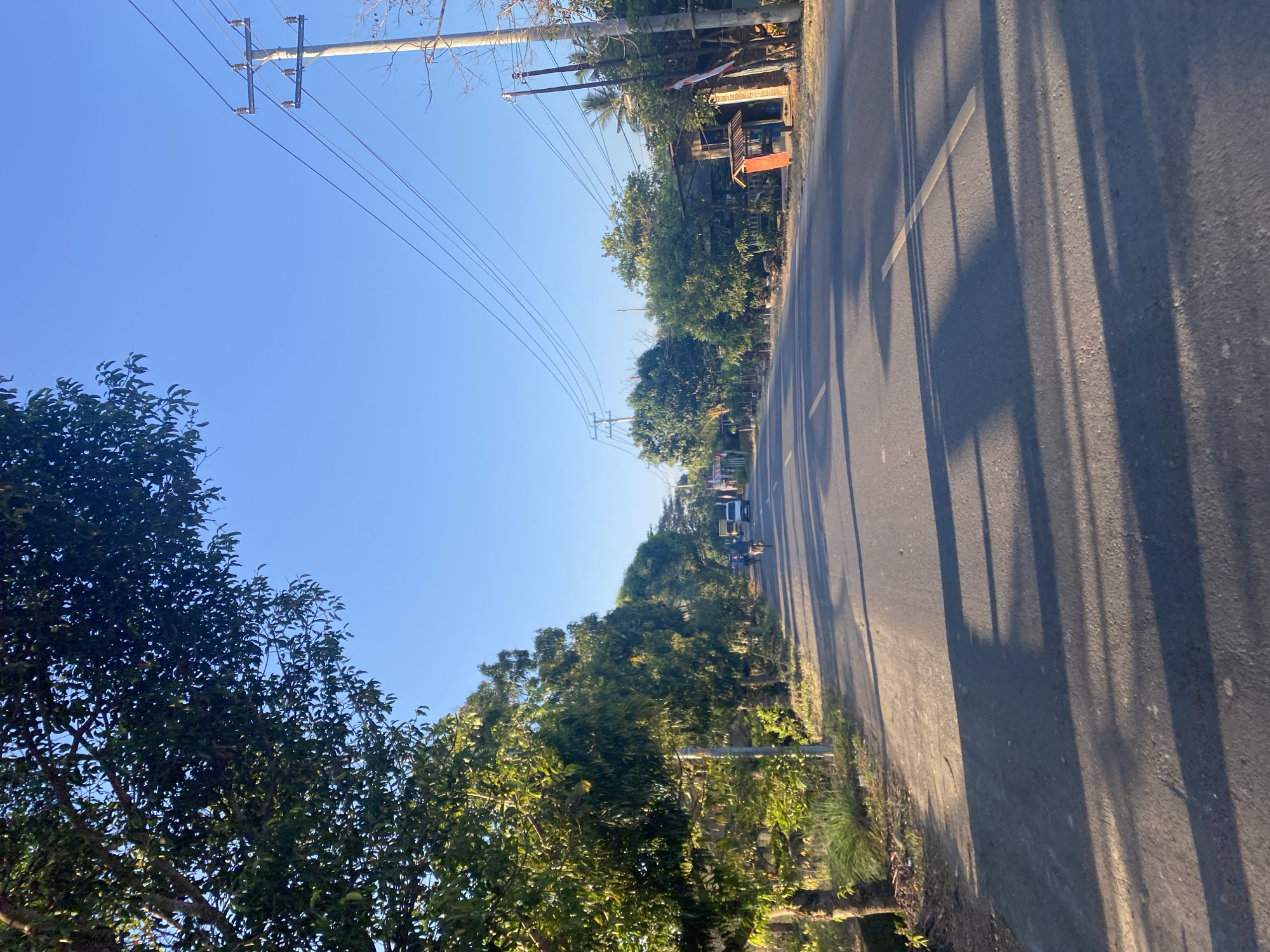 The image shows a paved road running through a semi-rural area, likely in Tanah Harapan, Indonesia, given the hint. The road is lined with trees on both sides, with dense foliage creating a canopy in some areas. A few vehicles, including a white van and at least one motorcycle with a rider, are visible on the road, moving away from the viewer. Power lines are strung across poles on the right side of the road, and there are some simple structures and houses visible amongst the trees. The sky is clear and blue, indicating fair weather, and the sunlight casts long shadows across the road, suggesting it is either early morning or late afternoon. The overall impression is one of a quiet, everyday scene in a tropical or subtropical environment.
