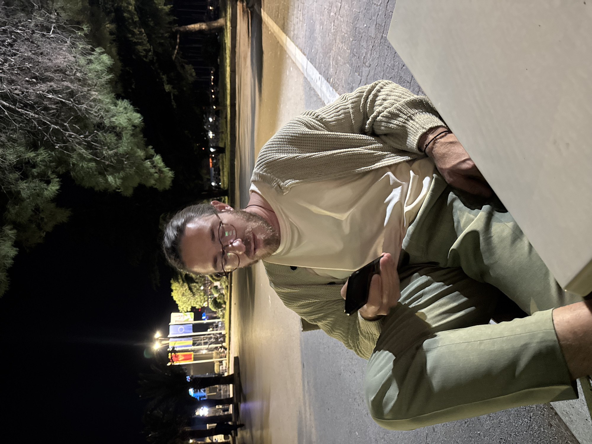 A man wearing glasses, a white t-shirt, a light-colored cardigan, and green pants sits outdoors at night. He is holding a black mobile phone in his right hand. Behind him, illuminated flags and lights indicate a public space in Bar, Montenegro. A large tree is visible to the left of the flags.