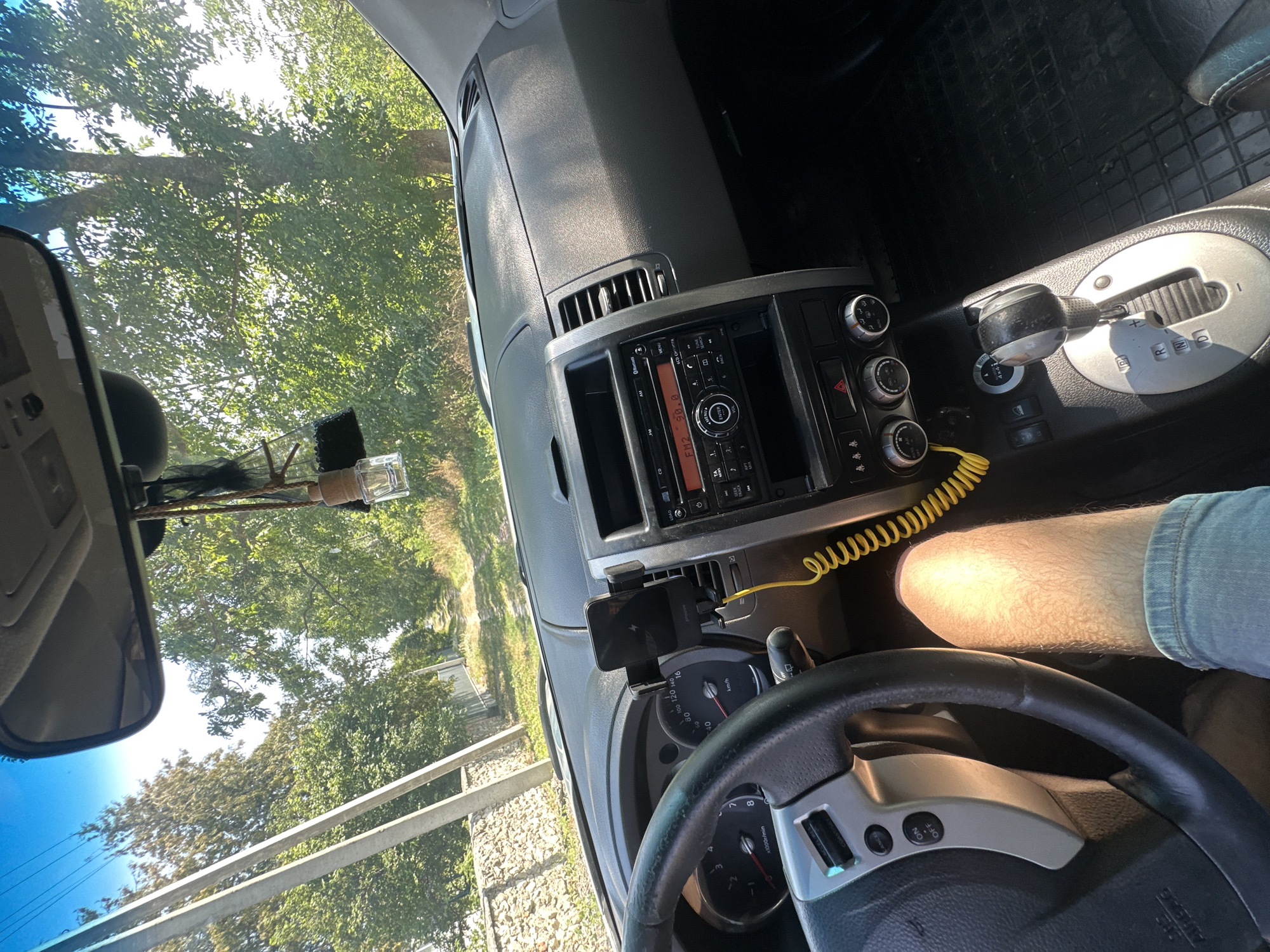 The image shows the interior of a car, likely a Nissan X-Trail, viewed from the driver's seat. The dashboard, steering wheel, gear shift, and radio are visible. A smartphone is mounted on the dashboard. Through the windshield, a dirt road lined with trees and a stone fence is visible, suggesting a rural or suburban setting. The sky is clear and blue. The leg of a person wearing denim shorts is visible in the lower right corner. The car appears to be parked or moving slowly. The location is identified as Makiv, Ukraine.