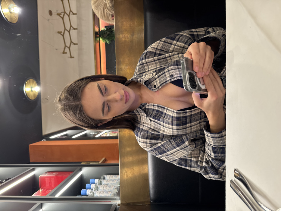 An image captures a young adult female seated in a black booth at what appears to be a restaurant or bar in Verona, Italy. She has long, straight dark brown hair with a middle part and is looking down intently at a smartphone held in both hands. She wears an unbuttoned blue and beige plaid shirt over a dark-colored bra or top. On the white table in the foreground, silverware, including a fork and knife, is visible on the left. The background features a golden-hued partition, above which are dark shelves with linear LED lighting illuminating rows of bottles with blue caps and red boxes. The ceiling is dark and reflective, showing reflections of circular brass light fixtures and a larger, hexagonal-patterned chandelier. A partial view of light-colored hair from another patron is visible in the far-right reflection.