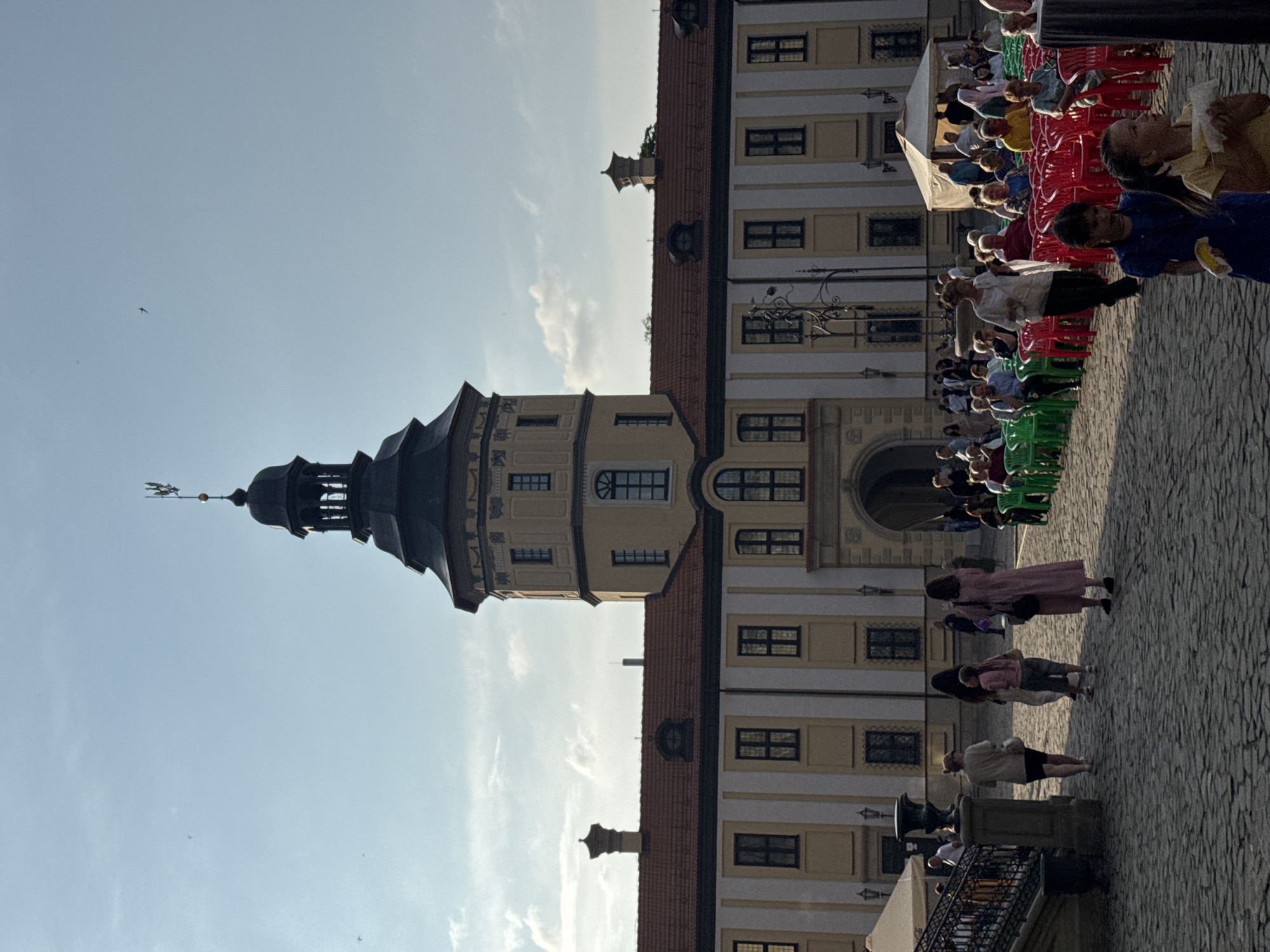 The image shows a crowd gathered in the courtyard of Nesvizh Castle in Nesvizh, Belarus. The castle, a UNESCO World Heritage site, is a large, yellow building with a prominent tower topped with a weather vane. In front of the castle, rows of red and green plastic chairs are set up, suggesting an outdoor event or performance. People of various ages are seated in the chairs or standing in the courtyard. Some individuals are walking in the foreground, while others are gathered near tents set up to the right of the castle. The courtyard is paved with cobblestones, and the sky is partly cloudy with a few birds visible. The lighting suggests it is late afternoon or early evening.