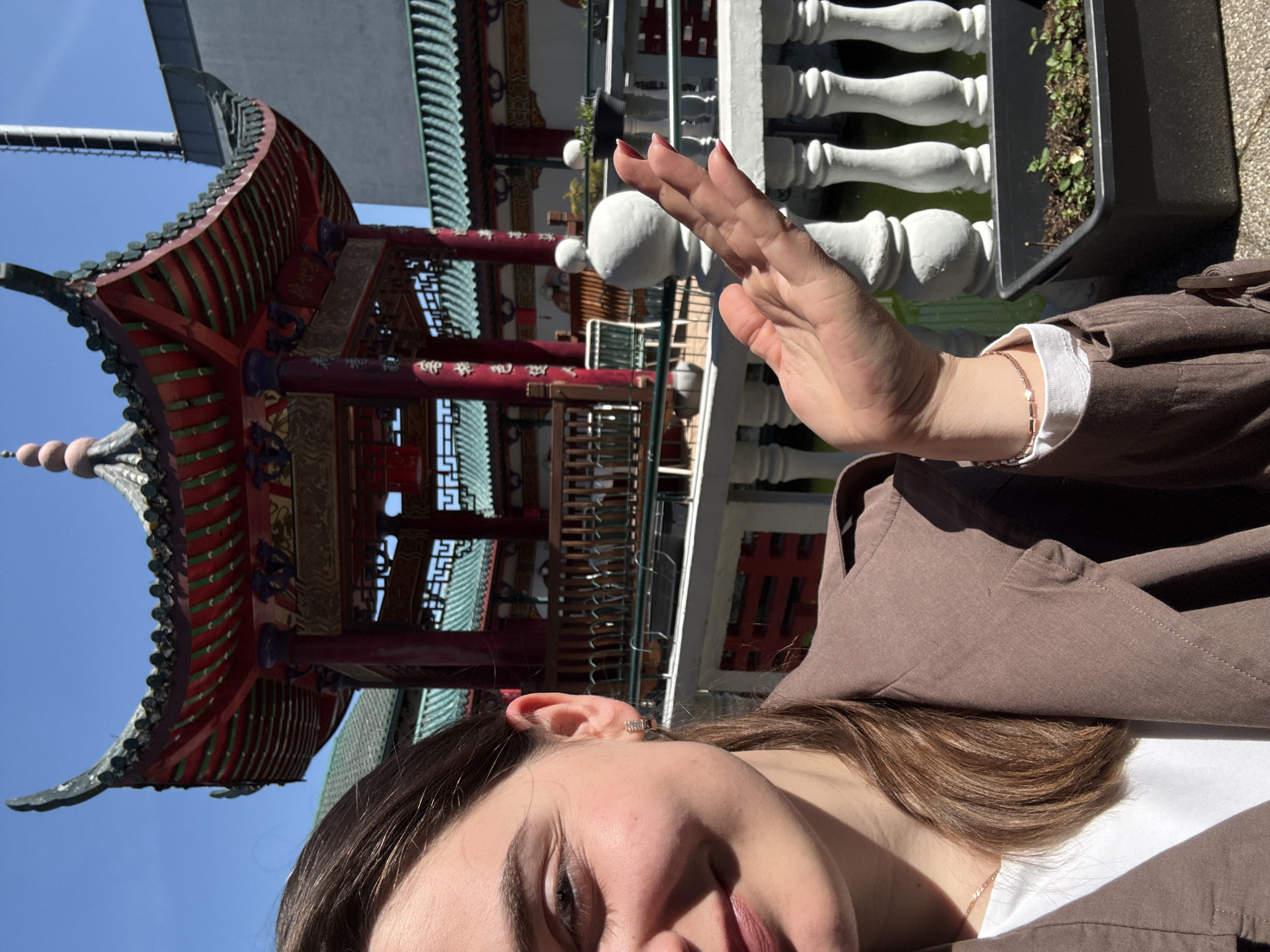 A brightly lit, outdoor selfie captures a person in the foreground against a backdrop of East Asian-style architecture. In the bottom right, the face of a young person with brown hair is partially visible, showing one eye, part of the nose, and lips, partially obscured by shadows. They wear a brown jacket over a white top and a delicate gold-colored bracelet on their right wrist. Their right hand is raised, possibly shielding their face from the sun or making a gesture, with light pink nail polish visible on the fingernails. A small earring adorns their right ear.
The background is dominated by a distinctive pagoda-style structure, featuring multiple tiers of curved, dark-tiled roofs. The building is adorned with vibrant red and green painted wooden elements, pillars, and intricate decorative patterns. In the midground, a white balustrade with spherical embellishments runs horizontally, and a black rectangular planter containing small green plants is visible.
The sky is clear and blue, indicating bright, sunny weather during the daytime, likely mid-morning or afternoon due to the strong sunlight and shadows. The architecture suggests a cultural garden, temple, or similar feature. The image was taken in Hainburg an der Donau, Austria. No visible text is present.