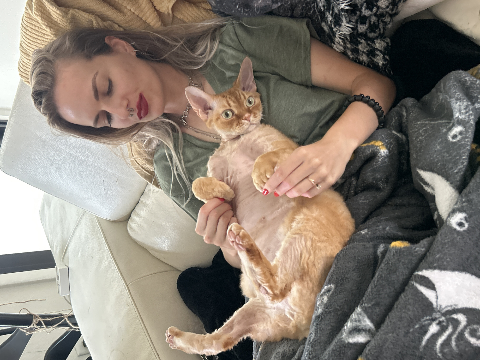 A person with blonde hair is lying on a white couch, holding an orange cat on its back. The person is wearing a green shirt and has a nose ring. The cat is looking directly at the camera. A gray blanket is covering the person and the cat. The location is indoors. The city is Cape Coral, United States of America.