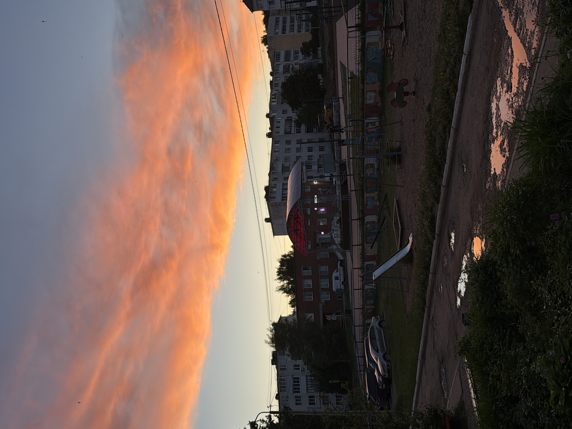 The image presents a serene urban or suburban landscape, likely in Noviy, Russia, captured during either sunrise or sunset, given the dramatic lighting. The sky is a dominant feature, with a broad swathe of vibrant orange and pink clouds occupying the right side, gradually transitioning to a lighter blue-grey on the left. Fine power lines stretch horizontally across the middle of the sky.
In the foreground, a dirt or unpaved road is visible, dotted with puddles that reflect the fiery hues of the sky, indicating recent rainfall. Green grass and bushes line the right side of this path. To the left of the path is a playground area, equipped with various elements including a red and green rocking horse, metal climbing structures, and a slide. A colorfully painted fence runs alongside the playground.
Beyond the playground and road, a row of multi-story apartment buildings, characteristic of Soviet-era architecture, lines the horizon. Trees are scattered among and in front of these buildings. A distinctive structure with a red, arched canopy or roof, illuminated by lights, is visible in the mid-ground. Several cars are parked on what appears to be a street or parking lot near the buildings. Faint, indistinct text, including "M-280", can be seen on a structure or sign in the mid-ground. No people are visible, and no specific activity is taking place, contributing to a quiet, contemplative atmosphere.