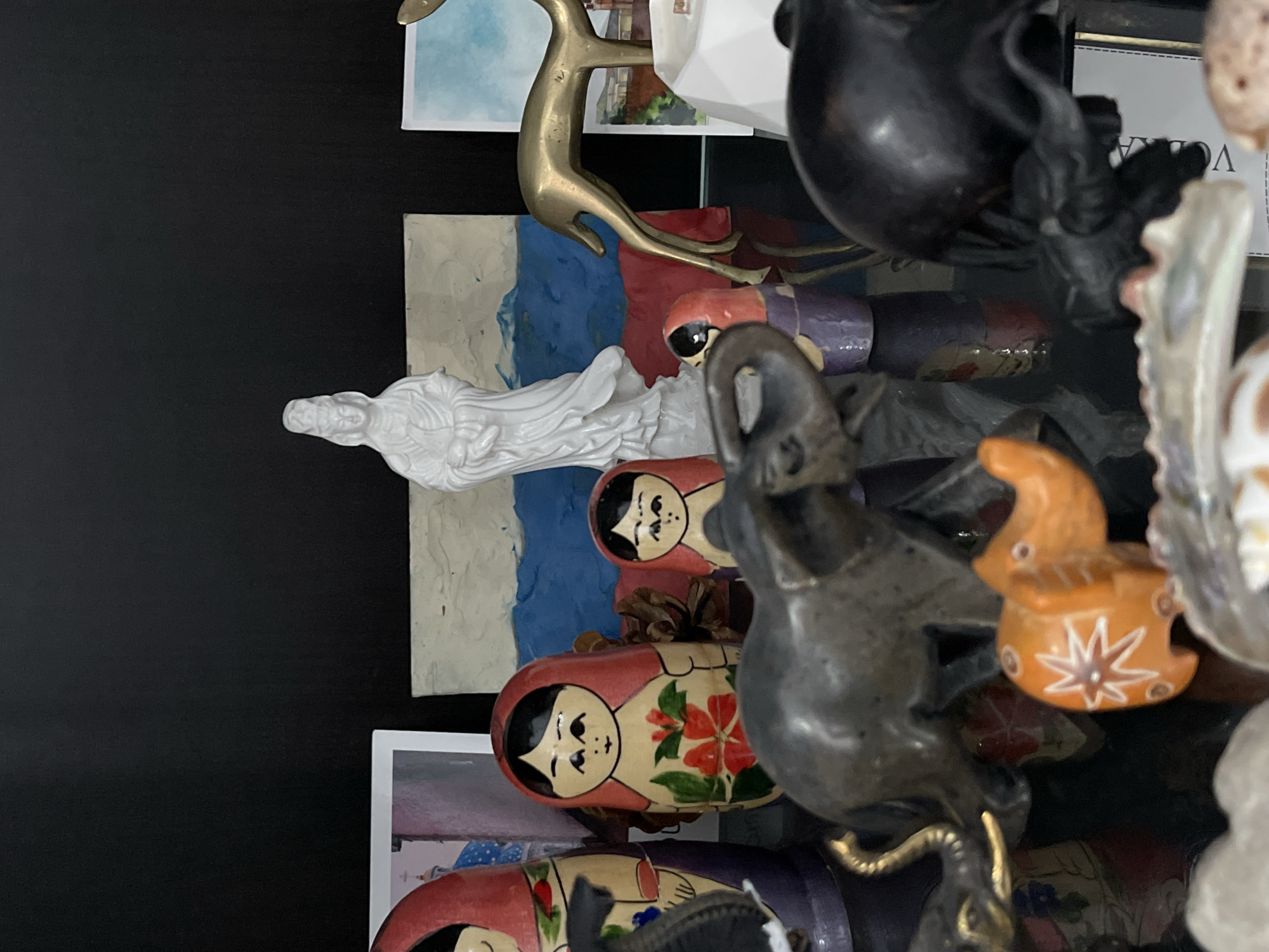 The image presents a cluttered display of objects against a dark background, likely a shelf or surface. Several items are identifiable: nesting dolls (Matryoshka dolls) with floral and facial designs, a white figurine of a person in flowing robes, a brass deer figurine, and a dark-colored statue of an elephant. Other items include what seems to be an orange decorative object with a star design and a dark figurine. There are postcards visible in the background. The scene seems to be of a random collection of items, with no clear indication of a specific location in Stavropol, Russia.