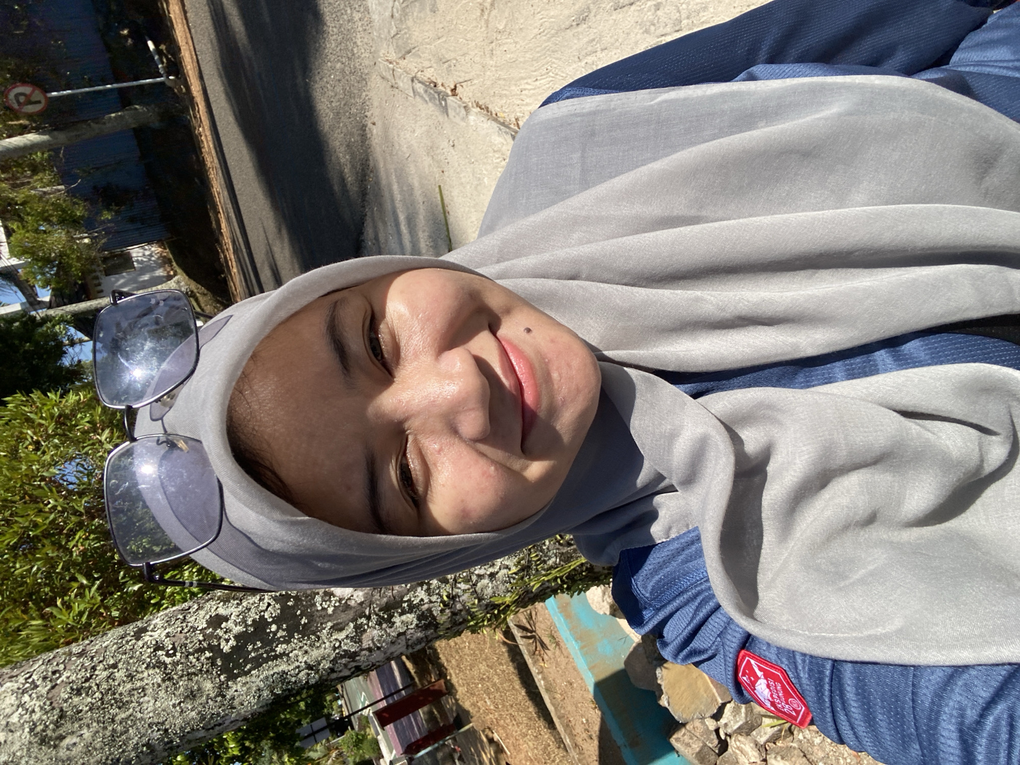 A young woman wearing a grey hijab and blue athletic shirt is captured in a selfie taken outdoors in Banjarbaru, Indonesia. She has sunglasses perched on top of her head and is smiling at the camera. The background shows a tree with textured bark, a paved road, and some vegetation, suggesting a park or roadside environment. The lighting indicates it is a sunny day. A small red patch with white lettering is visible on her shirt.