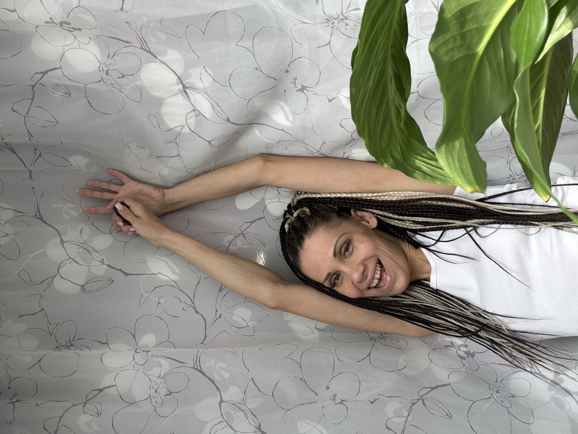 A woman with braided hair is lying on a bed with a floral patterned sheet. She is wearing a white t-shirt and has her arms extended. A large green plant is situated next to her. The image appears to have been taken indoors. There is no indication that this image was taken in Rusanovo, Russia.