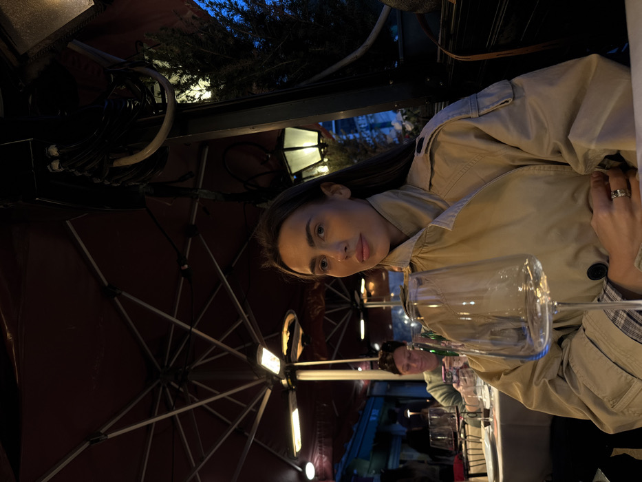 The image displays an outdoor dining scene in Venice, Italy, at night. The central subject is a woman, appearing to be in her 20s or 30s, with long dark hair and fair skin, positioned in the mid-ground. She wears a light beige trench coat over a plaid shirt and looks slightly to the viewer's right with a neutral expression. Her right hand, adorned with a multi-band ring on the ring finger, rests on a white tablecloth. In the immediate foreground, a clear, empty stemmed wine glass stands prominently.
In the blurred background, another individual, identified as a woman, is seated at a separate table also covered with a white tablecloth. This person is engrossed in a smartphone, holding it with both hands and looking down.
The environment is an outdoor restaurant patio covered by a large, dark red umbrella canopy, from which two active overhead heaters emit a warm orange glow. Overhead, bundles of black electrical wires are visible. To the right, a traditional-style street lamp with a glowing white diffuser illuminates a dark green artificial plant. The background indicates a dark blue evening sky, suggesting late dusk or night.
