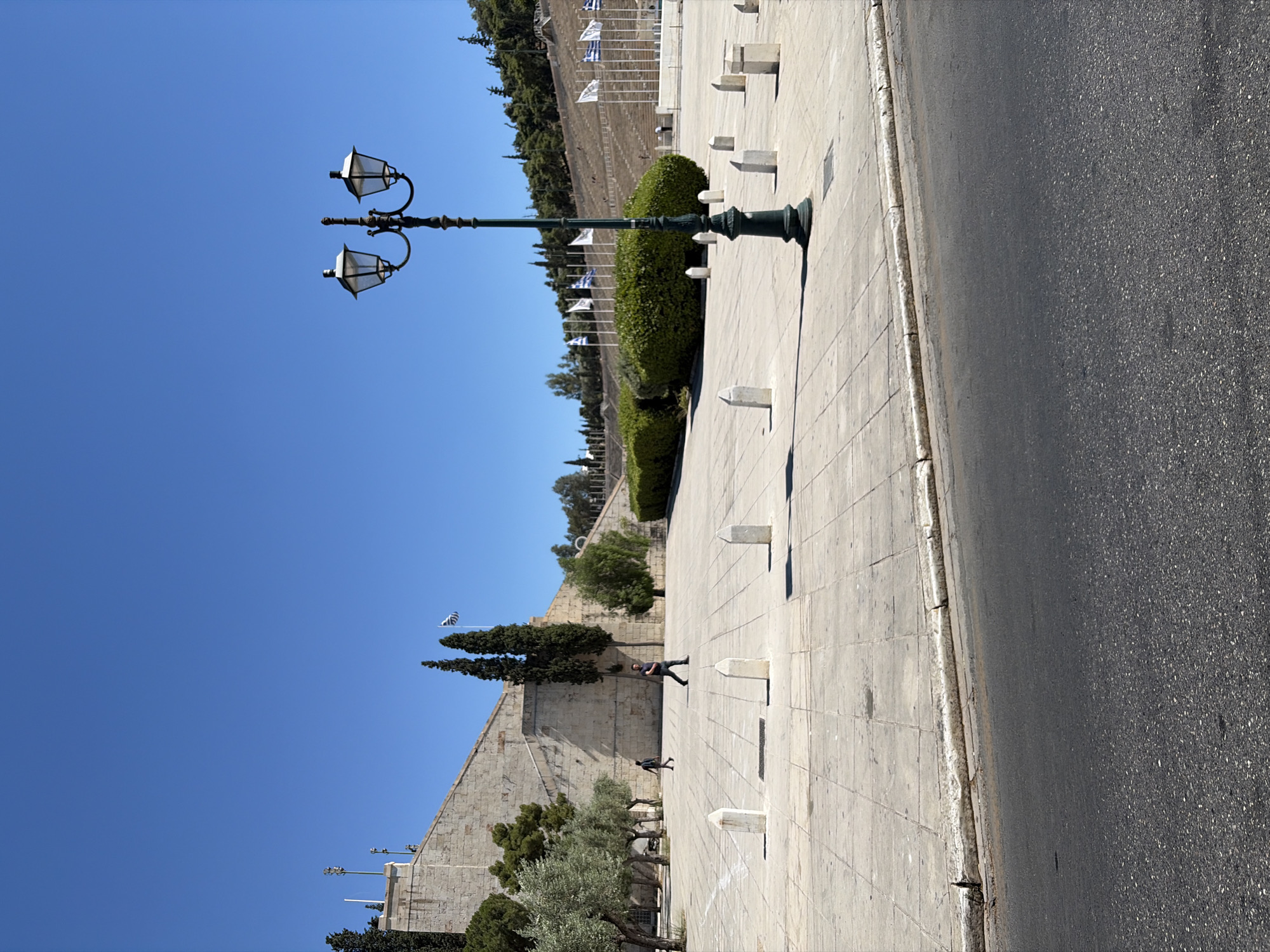This image depicts a view of the Panathenaic Stadium in Athens, Greece. The stadium's stone seating area is visible in the background, lined with numerous flags. In the foreground, a paved plaza is dotted with white bollards. A dark green lamppost with two lanterns stands prominently in the mid-ground. A section of asphalt road with a curb is visible in the bottom of the frame. Two individuals are walking across the plaza, and several olive trees and cypresses are scattered throughout the scene. The sky is clear and blue.