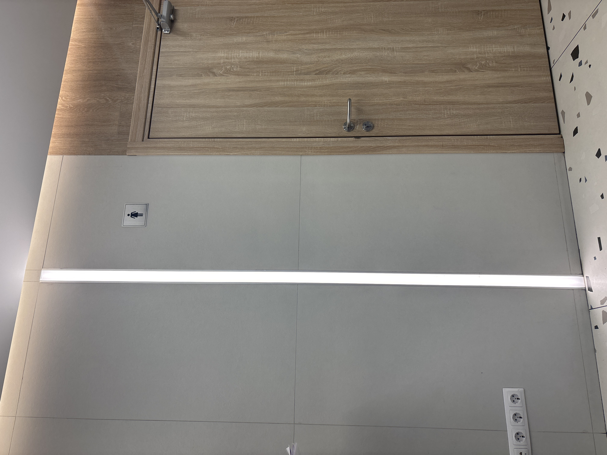 The image depicts an interior space, possibly a public restroom, in an unknown location. It features a wooden cabinet or paneling at the top, with a small metal handle. Below that is a gray tiled wall, divided into sections. A horizontal strip light illuminates the wall. A restroom sign with a female figure is on the left side of the wall. Three electrical outlets are stacked vertically on the lower right side of the wall. The environment is clean and modern. There are no people visible. The location in Saint Petersburg, Russia, cannot be definitively determined from the image alone.