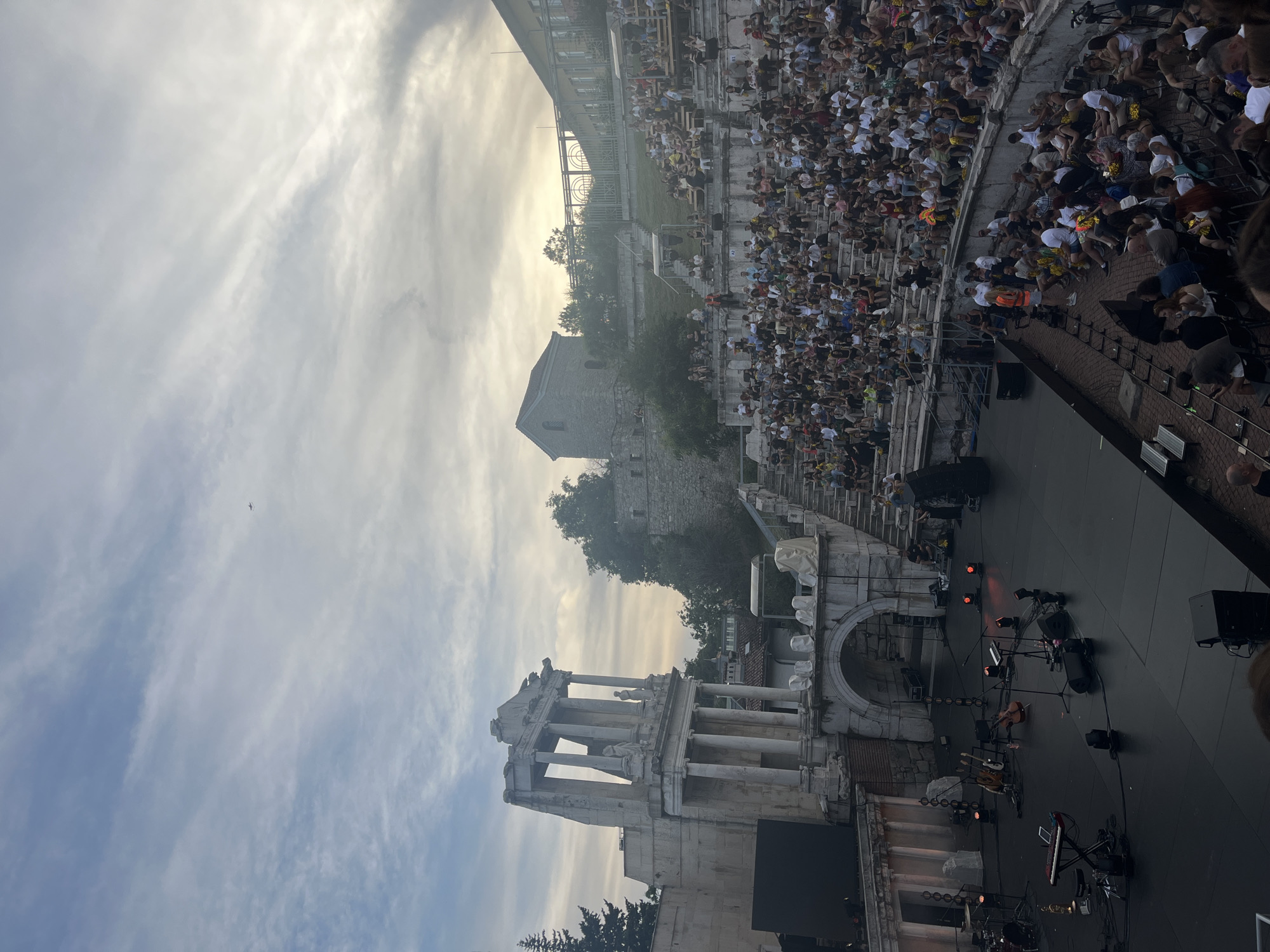An outdoor amphitheater, likely ancient, is filled with spectators at dusk, with a stage set up in the foreground. The amphitheater's stone seating curves upwards, accommodating a large crowd. On the stage, musical instruments and audio equipment are visible, suggesting a performance is about to take place or has recently concluded. To the left of the stage, a section of ancient ruins with columns and architectural detailing stands prominently against the sky. In the distance, a small airplane can be seen flying through the cloudy sky. The overall atmosphere appears to be one of anticipation and public gathering.