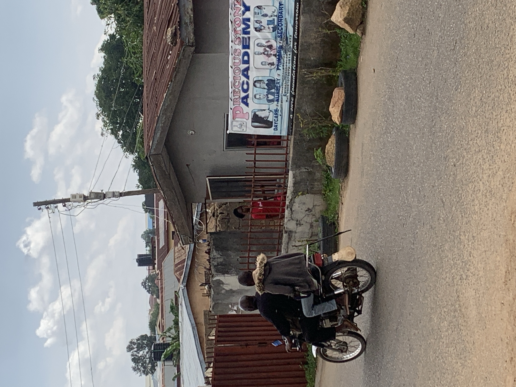 The image depicts a street scene in Jos, Nigeria, on a cloudy day. In the foreground, a motorcycle is carrying two individuals. The driver, an adult male wearing a black shirt, is at the front, while a passenger, possibly a younger male wearing a dark jacket with a fur-lined hood and blue jeans, sits behind. They are traveling away from the camera.
To the right of the motorcycle, a low-rise building with a corrugated metal roof is visible. A fence made of metal bars separates the street from the building's compound. Through the fence, a person wearing a red shirt can be seen standing inside the compound, near an open doorway. A banner is affixed to the exterior wall of the building. The banner advertises "PRECIOUS STONE ACADEMY" and lists "DAYCARE, NURSERY, PRIMARY, SECONDARY" education. It also includes contact information such as email and phone numbers, and the tagline "a sure foundation for tomorrow."
The environment appears somewhat urban with elements of a developing area. In the background, other buildings with tin roofs and some trees are present. Power lines are strung across the upper portion of the image, supported by utility poles. Some discarded tires filled with dirt and rocks are placed along the roadside in front of the building. The overall impression is of a functional, everyday street in a Nigerian city.