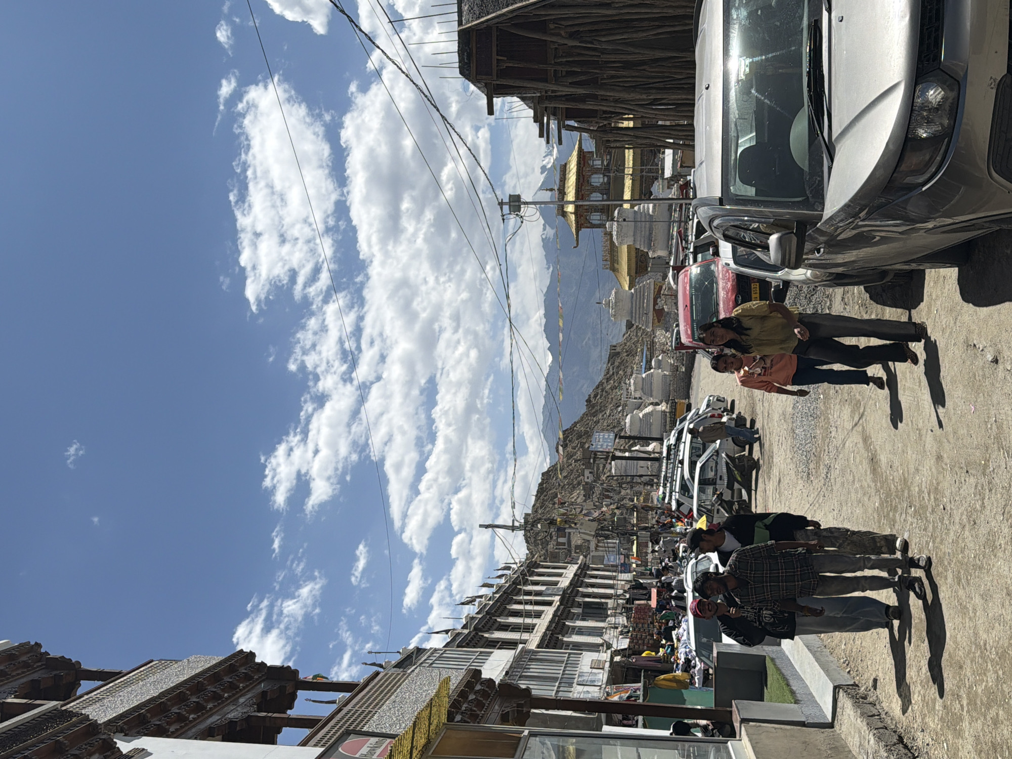 The photograph captures a bustling street scene in Leh, India, likely within the old town area, given the traditional architecture and mountainous backdrop. The time of day appears to be daytime, with clear blue skies dotted with fluffy white clouds.
In the foreground, a silver SUV is parked on the right side of the road, partially obscuring the view of other vehicles and pedestrians. To the left, a row of multi-story buildings lines the street, characterized by intricate wooden balconies and ornate carvings, typical of Leh's traditional style. Shops are visible at ground level, with goods displayed on the street.
Several people are walking along the street. Three men are in the left foreground, with one in the center wearing a checkered shirt. Two young women are walking on the right side of the street, further down, with one in a yellow top. In the background, more vehicles are parked, and a considerable number of people are seen browsing shops or walking.
The street is paved with rough dirt and gravel. Beyond the buildings and the street, the landscape rises to form rocky hills, characteristic of the Ladakh region. Snow-capped mountains are visible in the far distance, suggesting the high altitude of the location. Several white stupas, a common Buddhist religious monument, are scattered throughout the mid-ground. Prayer flags, strung between poles and buildings, flutter in the breeze, adding a touch of color to the scene. Electrical wires are strung across the sky, indicating the presence of infrastructure.
The overall impression is one of a vibrant, lived-in urban environment set against a dramatic natural landscape.