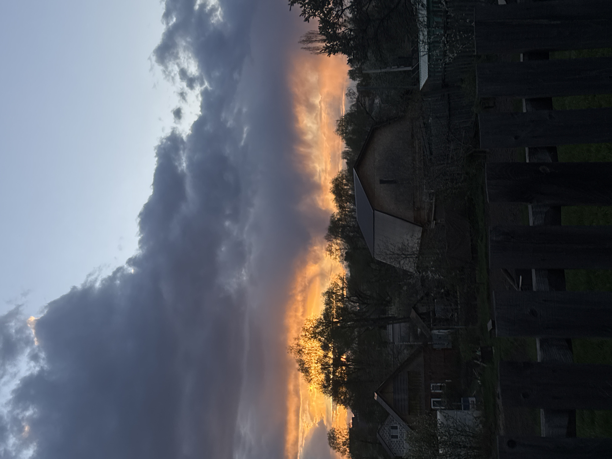 The image shows a sunset over a rural area, with a dark wooden fence in the foreground. Behind the fence, there are houses and trees silhouetted against the bright orange and yellow sky. The sky is partially covered with dark, ominous clouds. The location is likely in or near the city of Chaikovsky, Russia, based on the user's prompt. There are no people visible in the scene. The primary objects are the fence, houses, trees, and clouds. The main action or event is the sunset. The environment is a rural or suburban setting.