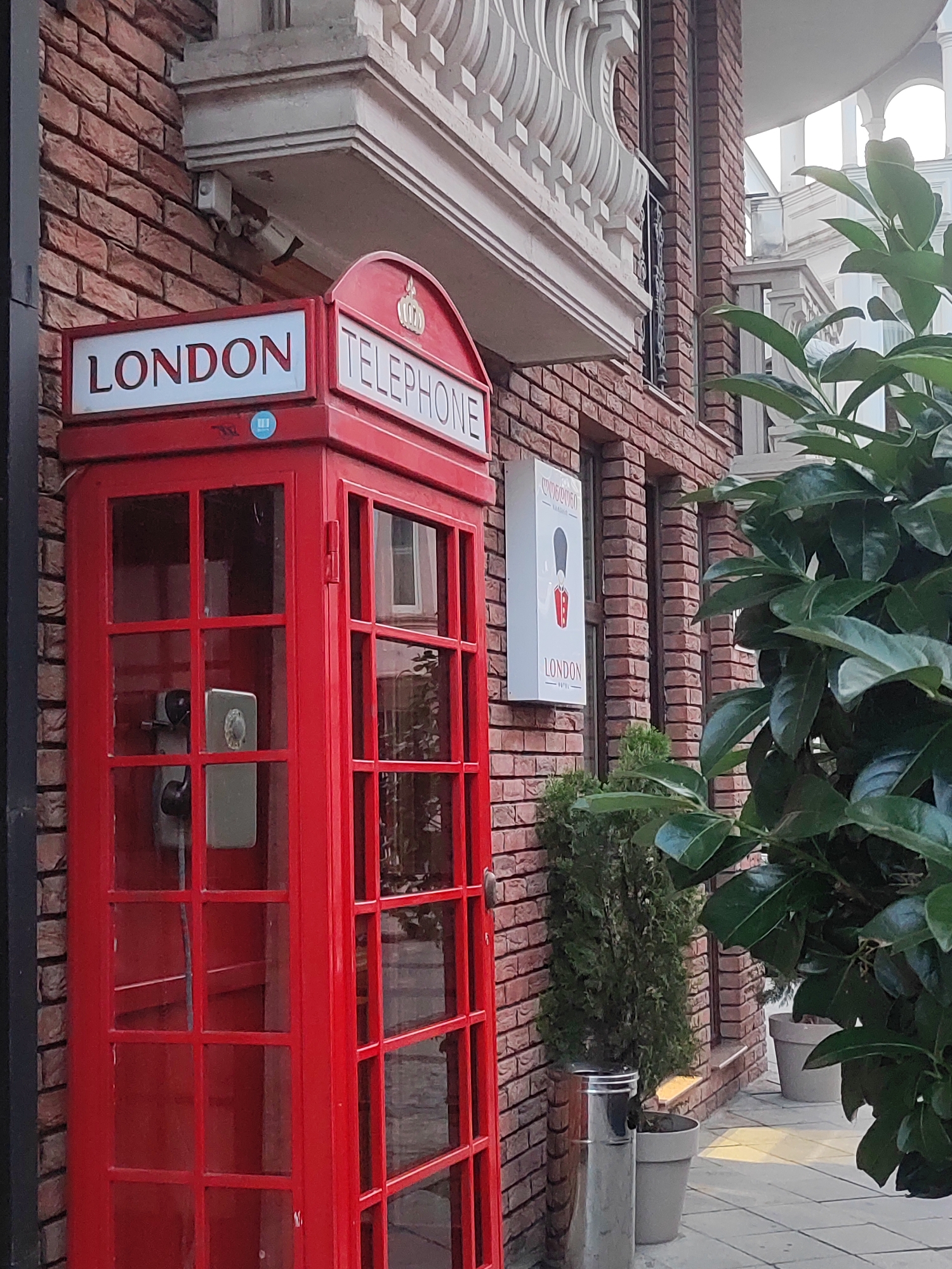 A red, classic British telephone booth stands in front of a brick building with white decorative elements and a balcony. A sign on the wall above the booth reads "LONDON TELEPHONE." To the right of the phone booth, another sign features a silhouette of a guard in a uniform and the word "LONDON." Green foliage partially obscures the view of the building. The scene is set in the city of Batumi, Georgia.