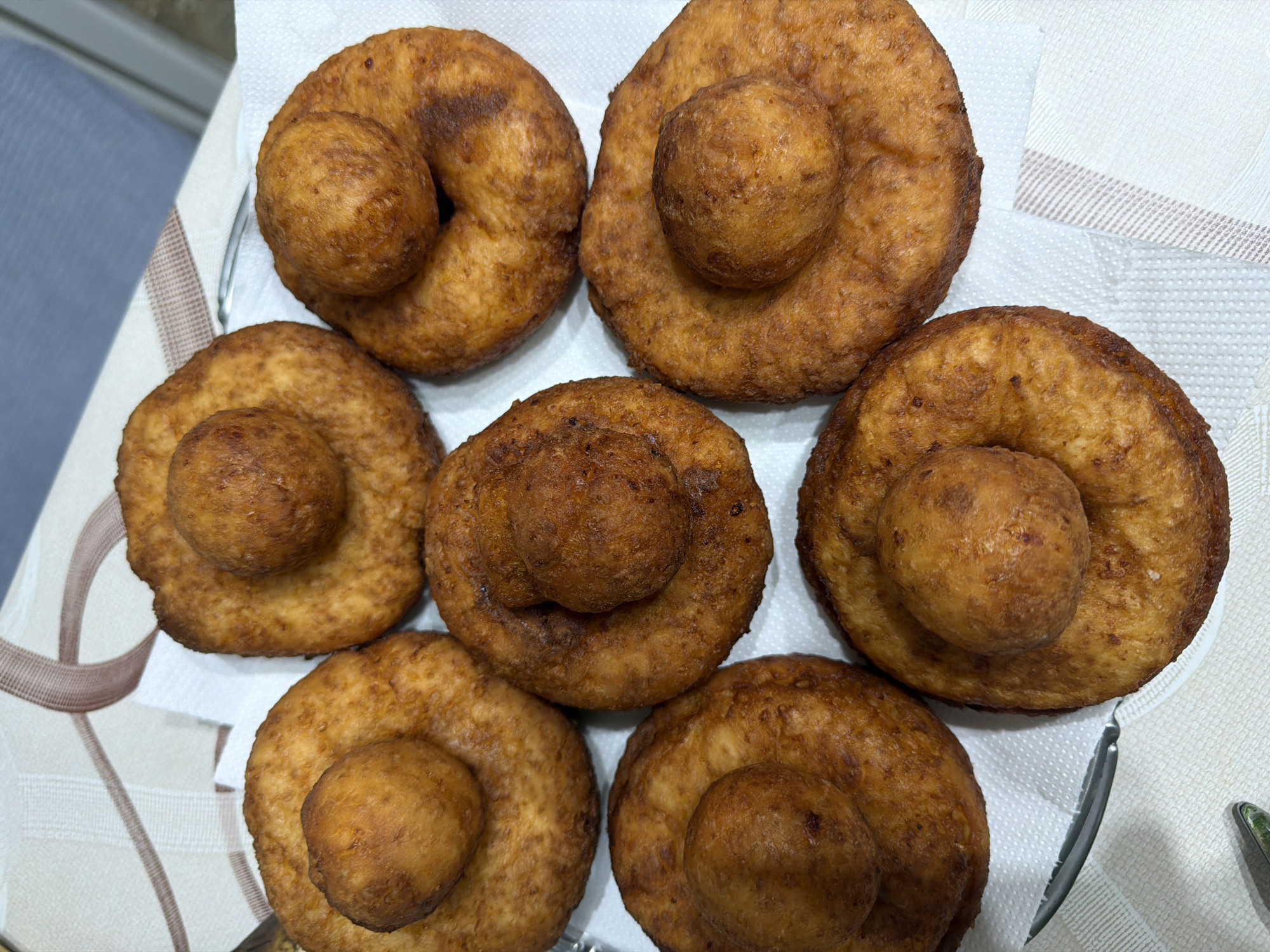 A top-down shot reveals seven golden-brown pastries arranged on a white surface that appears to be either a paper towel or a cloth, likely placed on a plate or tray. Each pastry consists of a ring-shaped base with a spherical component situated in the center. The surfaces of the pastries exhibit varied textures and colorations, with some areas appearing darker or more textured than others. The composition implies the pastries are homemade. The background is off-white. The location is Târgu Mureș, Romania is speculative.