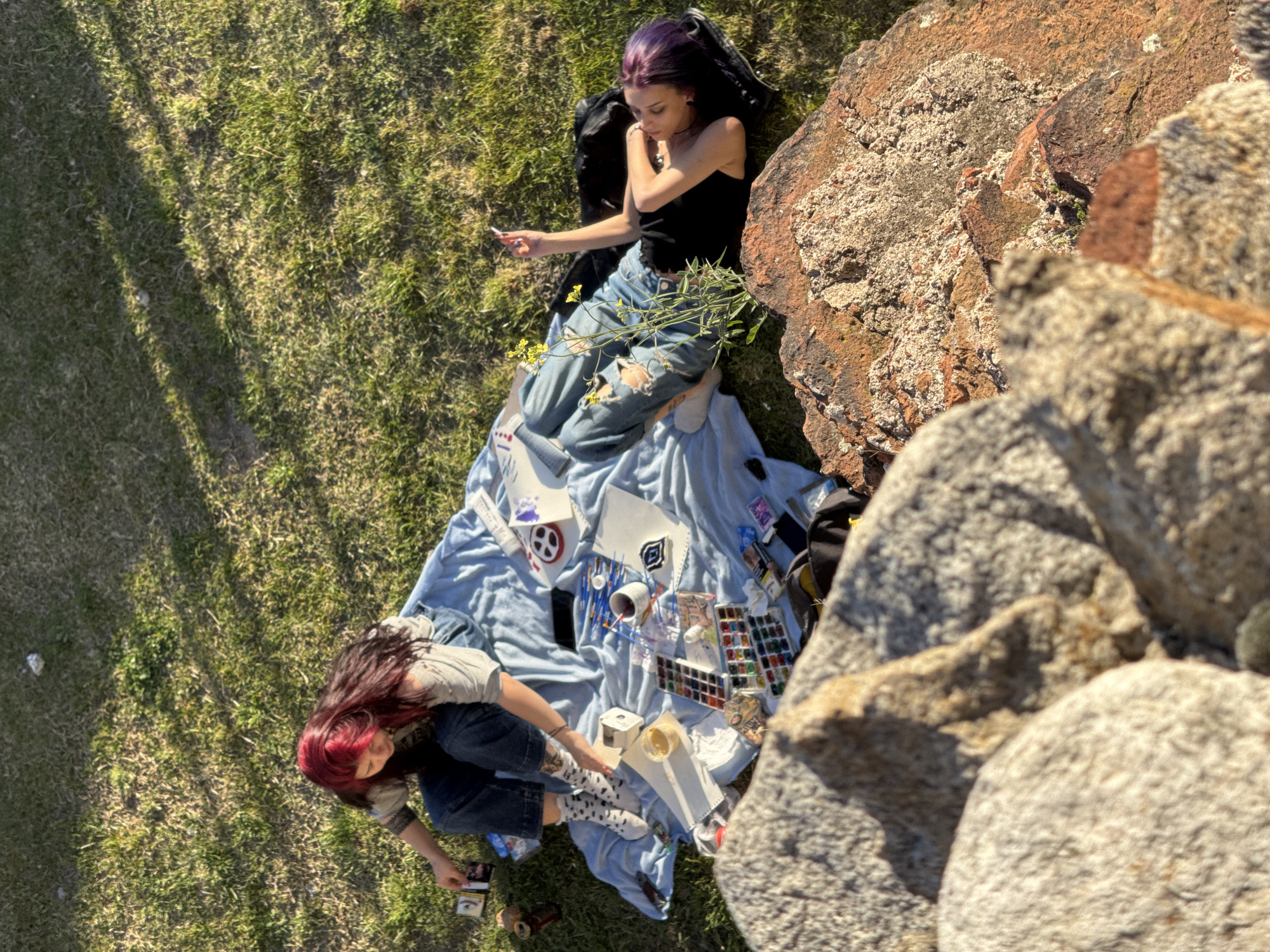 Two women are on a blanket on a grassy area next to rock formations. One woman, with purple hair and a black tank top, is lying down. The other woman, with red hair and a plaid shirt, is sitting and appears to be interacting with art supplies on the blanket. Art supplies, including watercolors, brushes, and paper, are scattered on the light blue blanket. The location may be Plovdiv, Bulgaria, but this cannot be determined from the visual data.