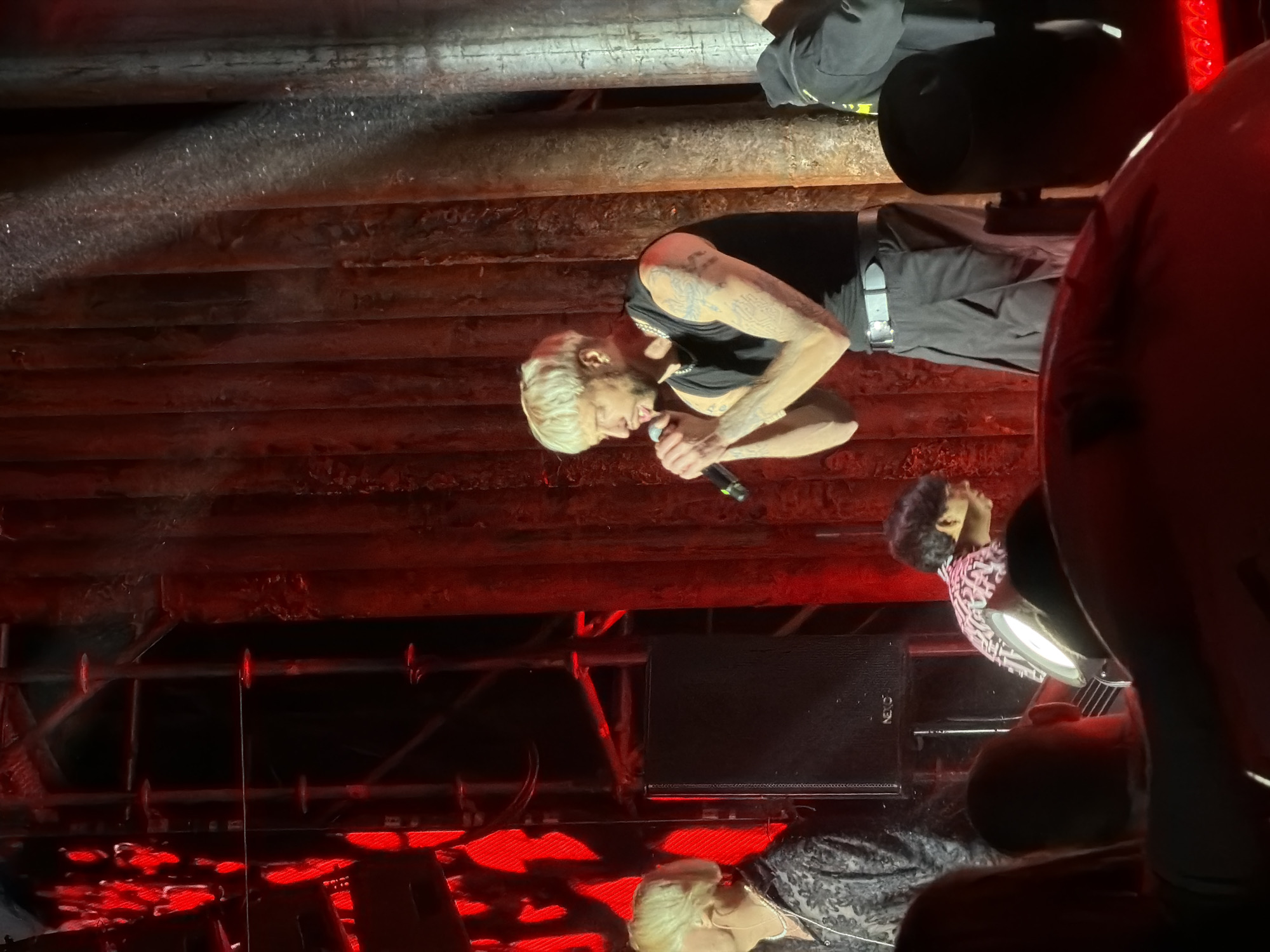 The image depicts a man with blond hair and tattoos on his arms, wearing a black tank top and dark pants, holding a microphone and singing on a stage. He is positioned in the center right of the frame. To his left, another person with blond hair and a patterned shirt is visible. In the background, red lighting illuminates a textured, vertical surface, possibly stage curtains or a backdrop. A speaker is mounted on the left side of the stage. The foreground is partially obscured by the heads and shoulders of audience members. There is no information in the image to indicate the location in Volzhsky, Russia.