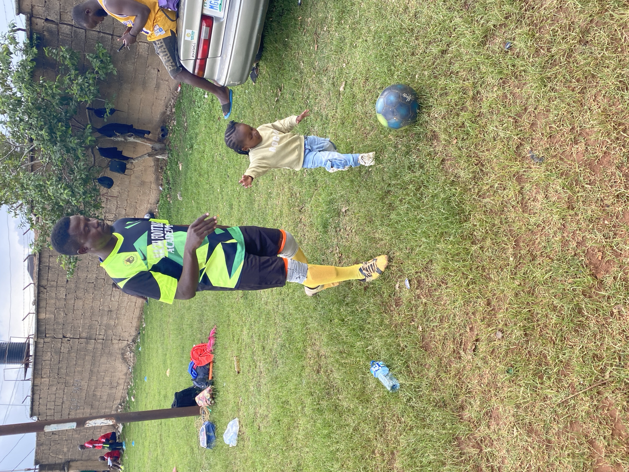 The image captures an outdoor scene at what appears to be a soccer practice or casual gathering in Gura Top, Nigeria, under bright daylight. The ground is a mix of green grass and patches of dirt.
In the foreground, an adult male with dark skin and short dark hair stands prominently, wearing a black and lime green soccer jersey with "FOOTBALL ACADEMY" text, black shorts, yellow socks, and soccer cleats. He holds his hands slightly up, as if gesturing or mid-instruction.
To his right, a young child with braided hair, dressed in a light yellow long-sleeved top (with visible text "ADGA" or similar) and light blue jeans, lies on their back on the grass. A worn blue and green soccer ball rests nearby.
In the background, another adult male in a yellow sleeveless top (resembling a Lakers jersey) and dark shorts leans against a light-colored Toyota sedan, looking at his phone. Dark clothing or bags hang from a small leafy tree growing against a weathered brick wall that forms the backdrop. The wall has barbed wire on top and a faint "CAR PARK" inscription. A partially visible license plate on the car reads "MG...".
Various items are scattered on the grass, including a plastic water bottle near the main adult, and colorful sports equipment like cones and bags in the far left background, where several other figures in red shirts are partially visible. A large dark water tank is also visible behind the wall. The bright, clear daylight suggests it's likely midday or afternoon. The scene conveys a relaxed yet active atmosphere, typical of community sports.