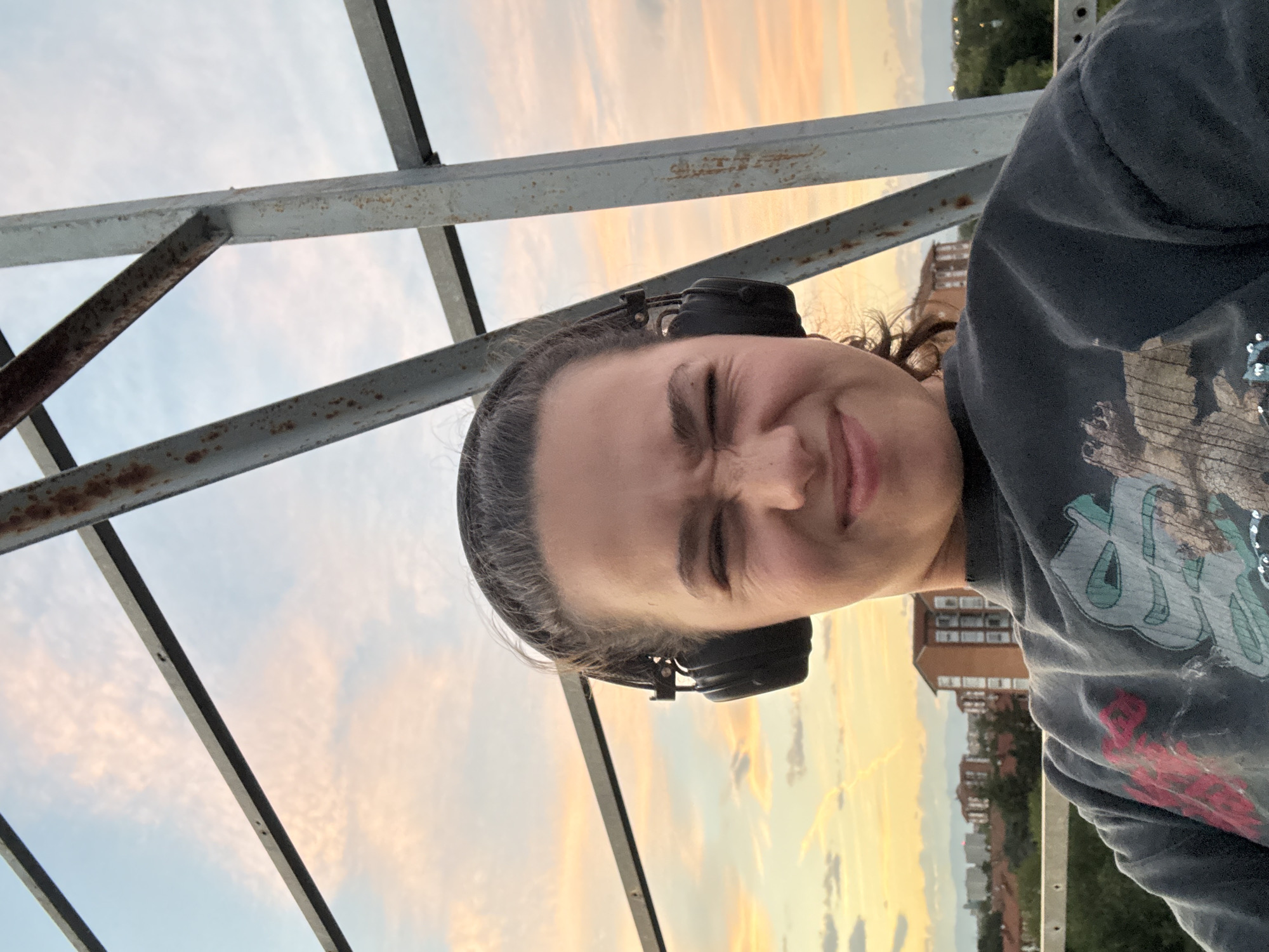 A young person with dark hair pulled back, wearing black headphones and a dark gray t-shirt with a graphic print, makes a face at the camera. They are standing on an elevated structure with rusty metal beams against a sky with pink and orange clouds at sunset. In the background, buildings and trees are visible, suggesting an urban environment. The scene appears to be in Milan, Italy, as indicated by the architecture and landscape.
