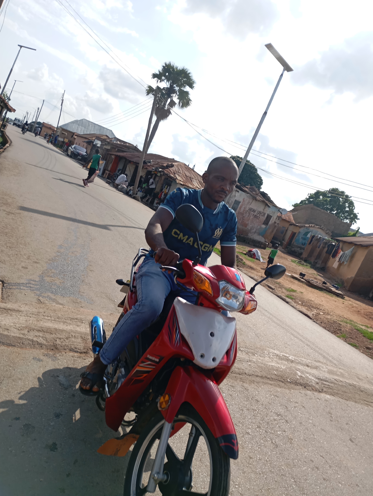 The image captures a young man seated on a red and white motorcycle, facing forward and looking directly at the camera. He is wearing a blue and white t-shirt with a logo that appears to be "CMAGNA" and blue jeans. His left foot is visible, wearing a black sandal. The motorcycle has a blue exhaust pipe. The scene appears to be a street in Shendam, Nigeria. The road is paved with asphalt and shows signs of wear and tear, with some loose gravel and patches of dirt. On either side of the road are a number of buildings with corrugated metal roofs, typical of residential structures in the area. Some of the buildings show signs of aging and disrepair, with faded paint and exposed materials. In the background, there are a few scattered trees, including a palm tree, and electrical wires crisscross overhead. The sky is partly cloudy with a bright, diffused light, suggesting it is daytime. Other people are visible in the background, including one person walking away from the camera in a green shirt and another group of individuals near the buildings. The overall impression is of a typical street scene in a Nigerian town.