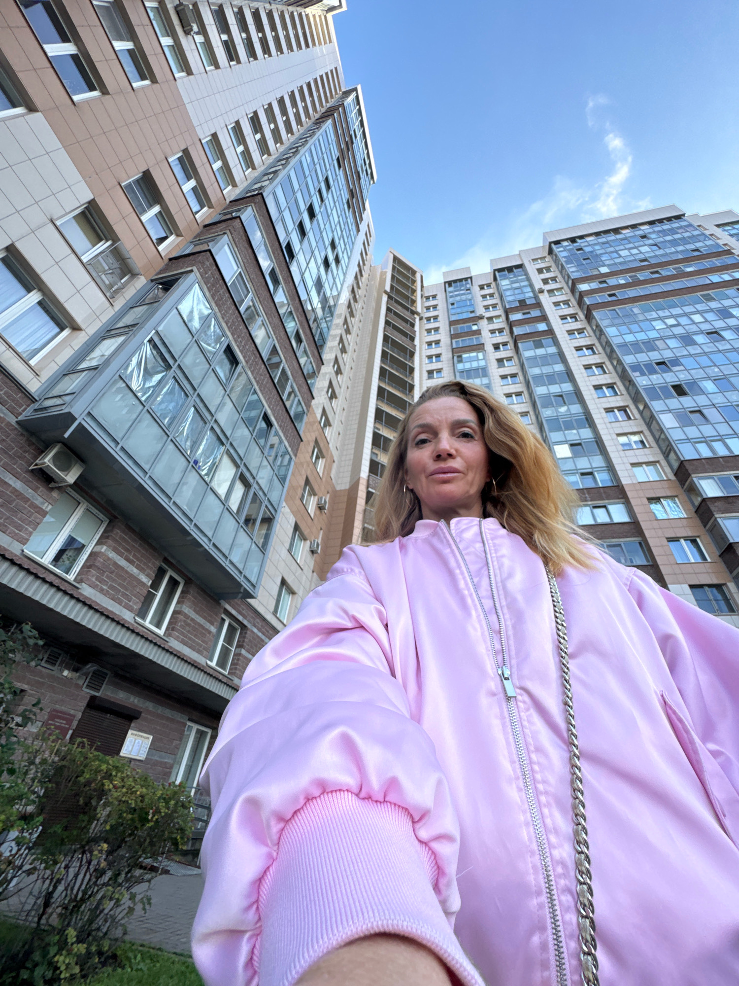 An outdoor, eye-level, medium-shot selfie shows a woman with blonde hair and fair skin standing in front of two tall apartment buildings. The woman is wearing a pink satin bomber jacket and a silver chain purse. The buildings are modern, with glass windows and balconies. The sky is blue with a few white clouds. The woman is looking at the camera and smiling. There is a bush in the foreground.