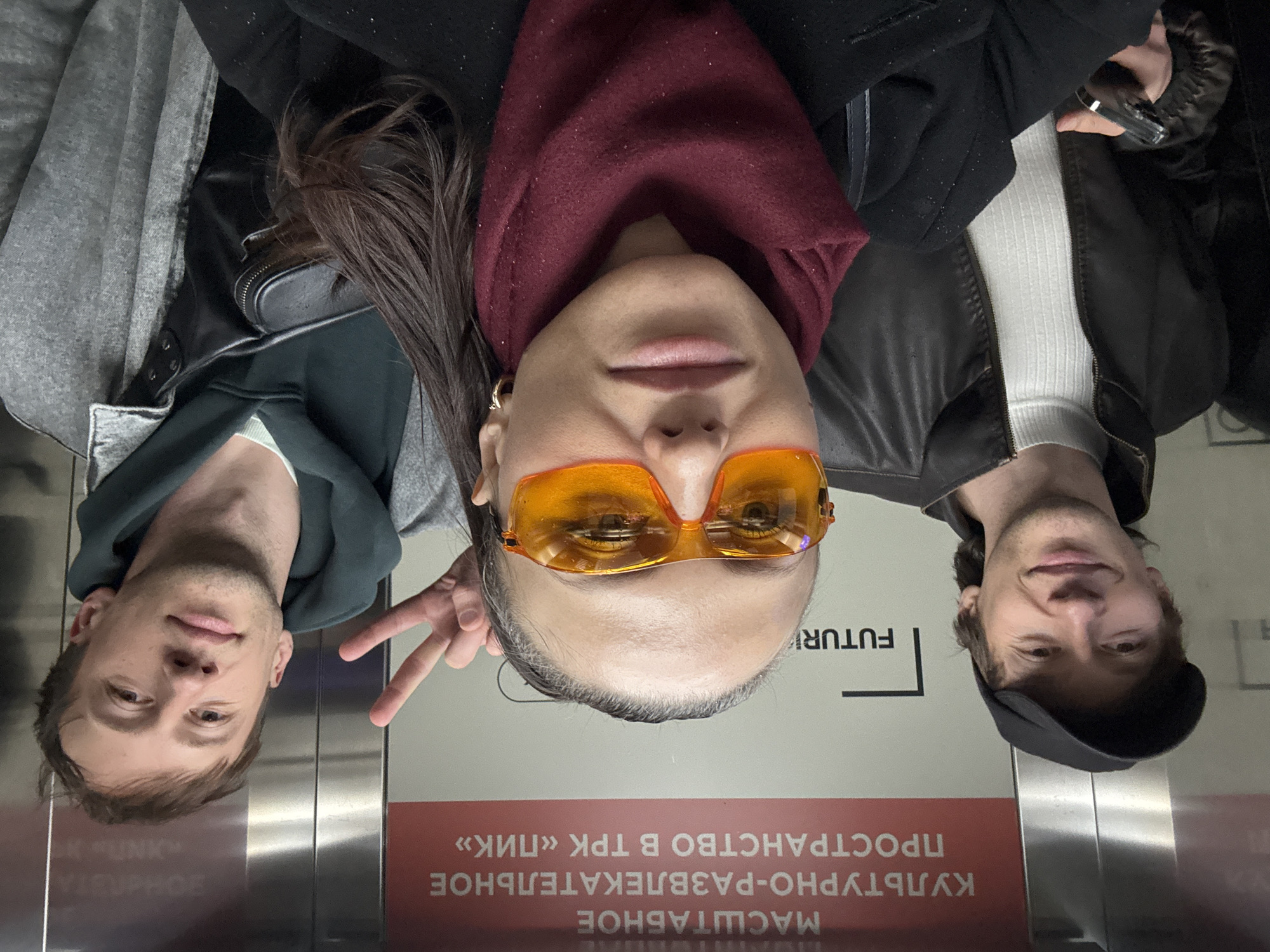 Three individuals are in an elevator in Saint Petersburg, Russia, taking a selfie. The elevator's interior features signage in Russian, including the phrase "КУЛЬТУРНО-РАЗВЛЕКАТЕЛЬНОЕ ПРОСТРАНСТВО В ТРК «ПИК»," which translates to "Cultural and entertainment space in TRK 'PIK'," indicating they are likely within the PIK shopping and entertainment complex.
The person in the center is wearing orange-tinted glasses and a maroon scarf. The individual on the left is making a peace sign. The individual on the right is wearing a black leather jacket over a white ribbed shirt and a black cap.