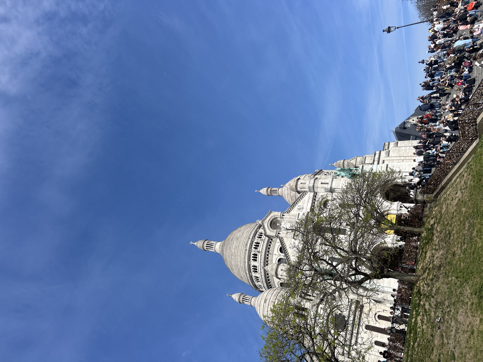 The Sacré-Cœur Basilica is shown on a bright day in Paris. The structure is mostly white and is capped by several domes and turrets. A bare tree partially obscures the front of the building. Many people are seated in the foreground on stairs and ledges. A lamppost is visible in the upper right. The sky is a clear, bright blue.