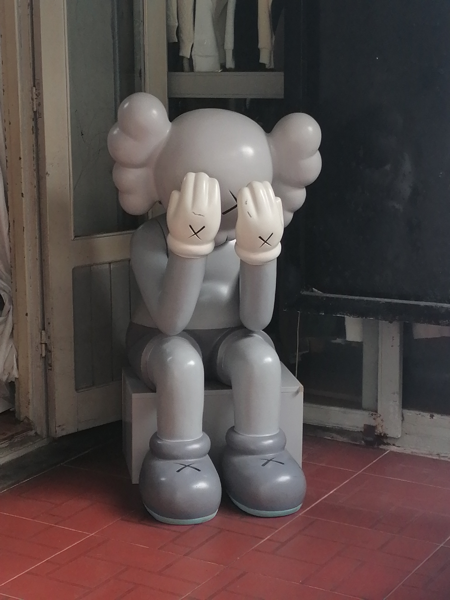 A grey KAWS sculpture sits on a cube-shaped platform on a red-tiled floor. The sculpture is seated, with its hands covering its eyes. The sculpture is located in Da Lat, Vietnam. A partially visible door frame is in the background on the left, and a dark, blurry surface is visible on the right. Additional objects are visible on a shelf in the background above the sculpture.