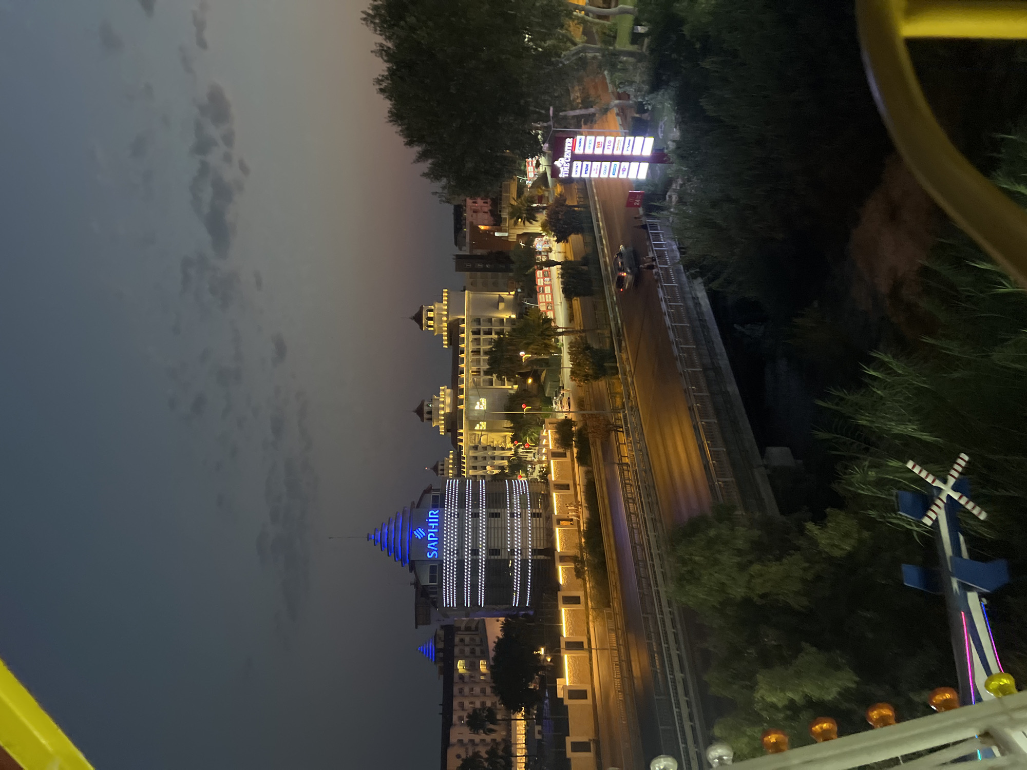 A high-angle, medium shot captures a nighttime cityscape viewed from what appears to be a Ferris wheel. In the foreground, parts of the Ferris wheel's structure, including yellow and white framing and illuminated bulbs, are visible. The mid-ground features a multi-lane road with vehicle headlights and taillights suggesting moderate traffic. A bridge crosses over a vegetated area or waterway below the road. Dominating the background are several large buildings, illuminated by exterior lights. One building, labeled "SAPHİR" in blue neon, has a distinctive conical roof and is decorated with horizontal lines of white lights. Other buildings display architectural styles with ornate roofs and numerous illuminated windows. The sky above is a dark, dusky blue with scattered clouds. The overall scene depicts an urban environment at night, likely a tourist or commercial district, with leisure activities suggested by the Ferris wheel and the brightly lit establishments.