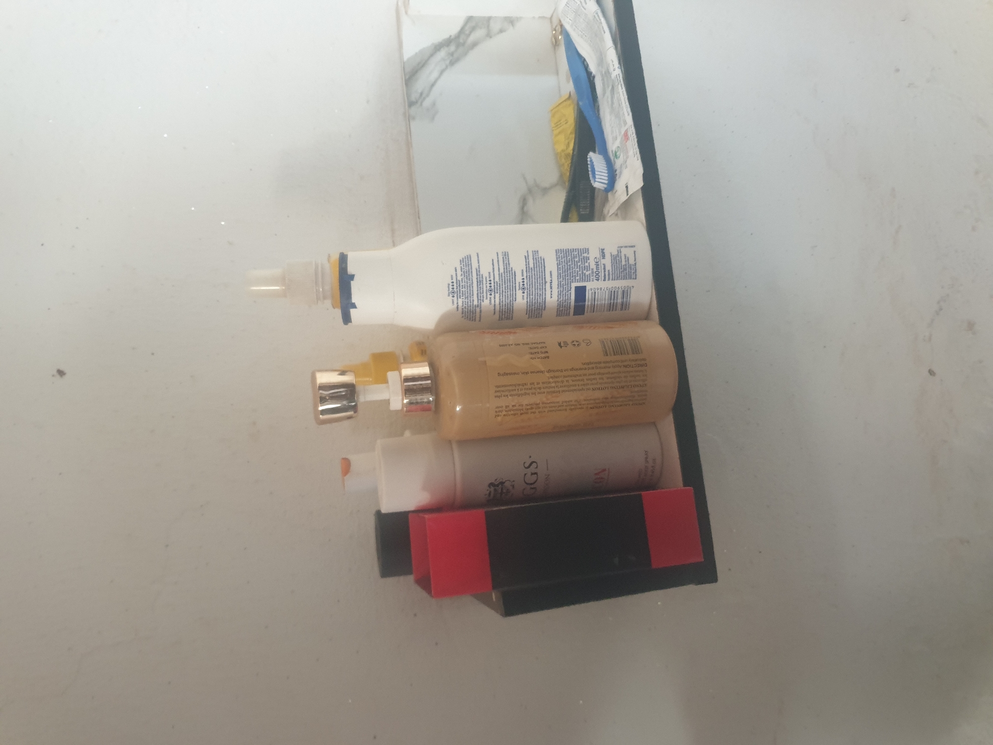 On a shelf attached to a white, textured wall, various toiletries are arranged. From left to right, a tall, black rectangular object with a red band is partially visible. Next to it is a white bottle with a silver cap. A tan-colored bottle with a gold cap stands behind it. Further to the right is a tall white bottle with a blue and yellow label, and a white pump dispenser. To the right of this, a blue toothbrush lies on a piece of paper. The background wall has a marbled pattern visible in the upper right. The lighting is ambient.