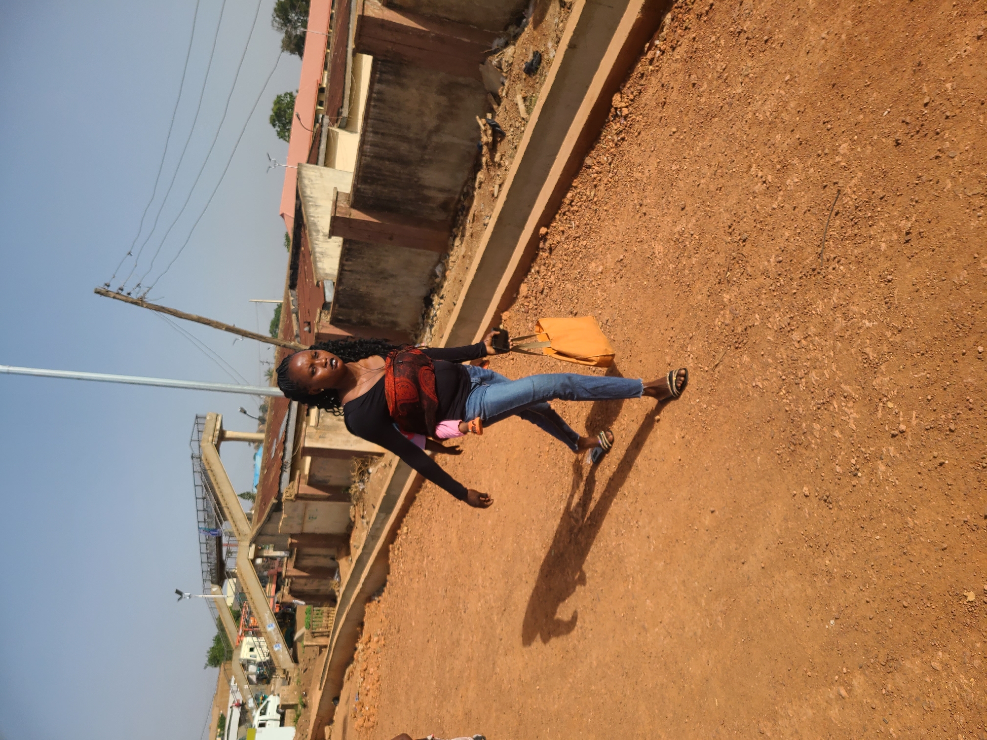 An outdoor scene depicts a woman standing on a dirt ground area in Gura Top, Nigeria. She wears a black long-sleeved shirt with a red floral pattern, blue jeans, and sandals. She carries a yellow tote bag and a pink object. The environment includes buildings, infrastructure like pedestrian bridges, and utility poles, suggesting an urban area. The sky is visible with a light blue hue.