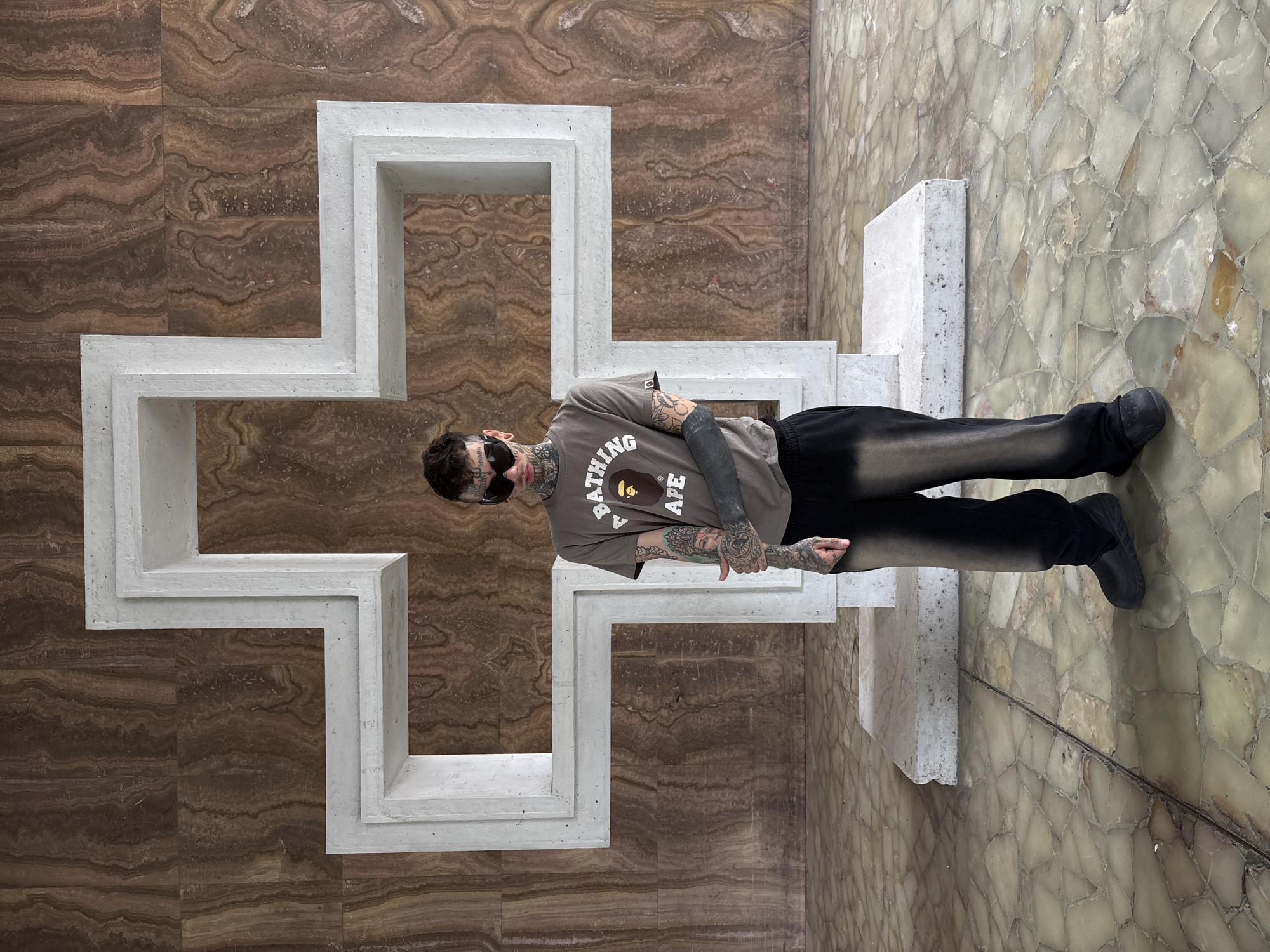 A solitary individual stands centered within an architectural interior, looking directly forward. The person wears black wrap-around sunglasses, a gray t-shirt with the "A BATHING APE" logo, dark, loose-fitting pants with a subtle vertical fade, and black slip-on shoes. Extensive dark tattoos are visible on their face, neck, arms, and hands. Their hands are positioned in front of their waist.
The background features a large wall composed of rectangular panels of horizontally veined brown and orange natural stone. Against this wall, a prominent, multi-tiered white concrete cross-like structure is fixed, showcasing a geometric, stepped design. The floor is paved with an irregular mosaic of light green, off-white, and amber translucent stone tiles.
This scene is located within the interior of the National Library of Argentina (Biblioteca Nacional Mariano Moreno) in Buenos Aires.