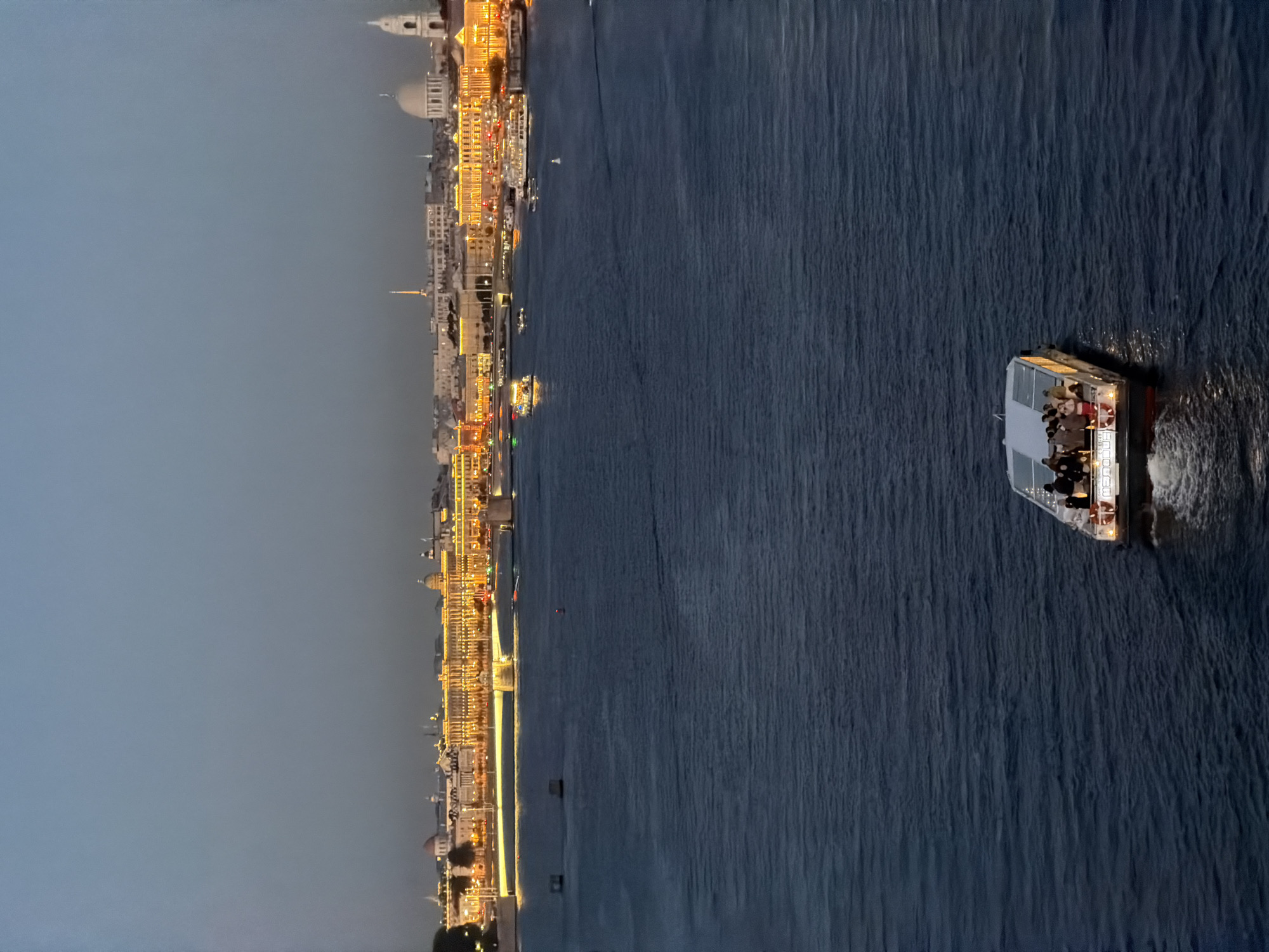 The image depicts a wide river at dusk, with the city skyline of Saint Petersburg, Russia, visible in the background. The sky is a deep twilight blue, transitioning to a lighter hue near the horizon. The city lights are illuminated, creating a warm glow along the waterfront. Numerous buildings, some with domes and spires, are identifiable, suggesting historical architecture. Several boats are present on the river. In the foreground, a smaller, modern boat with a glass enclosure is moving away from the viewer, carrying passengers visible on its deck. This boat is illuminated from within, casting a soft light on the water. Further in the distance, larger tour boats with their own lights are also navigating the river. The water itself is dark and choppy, reflecting the ambient light. The scene captures a serene evening ambiance in a prominent Russian city.