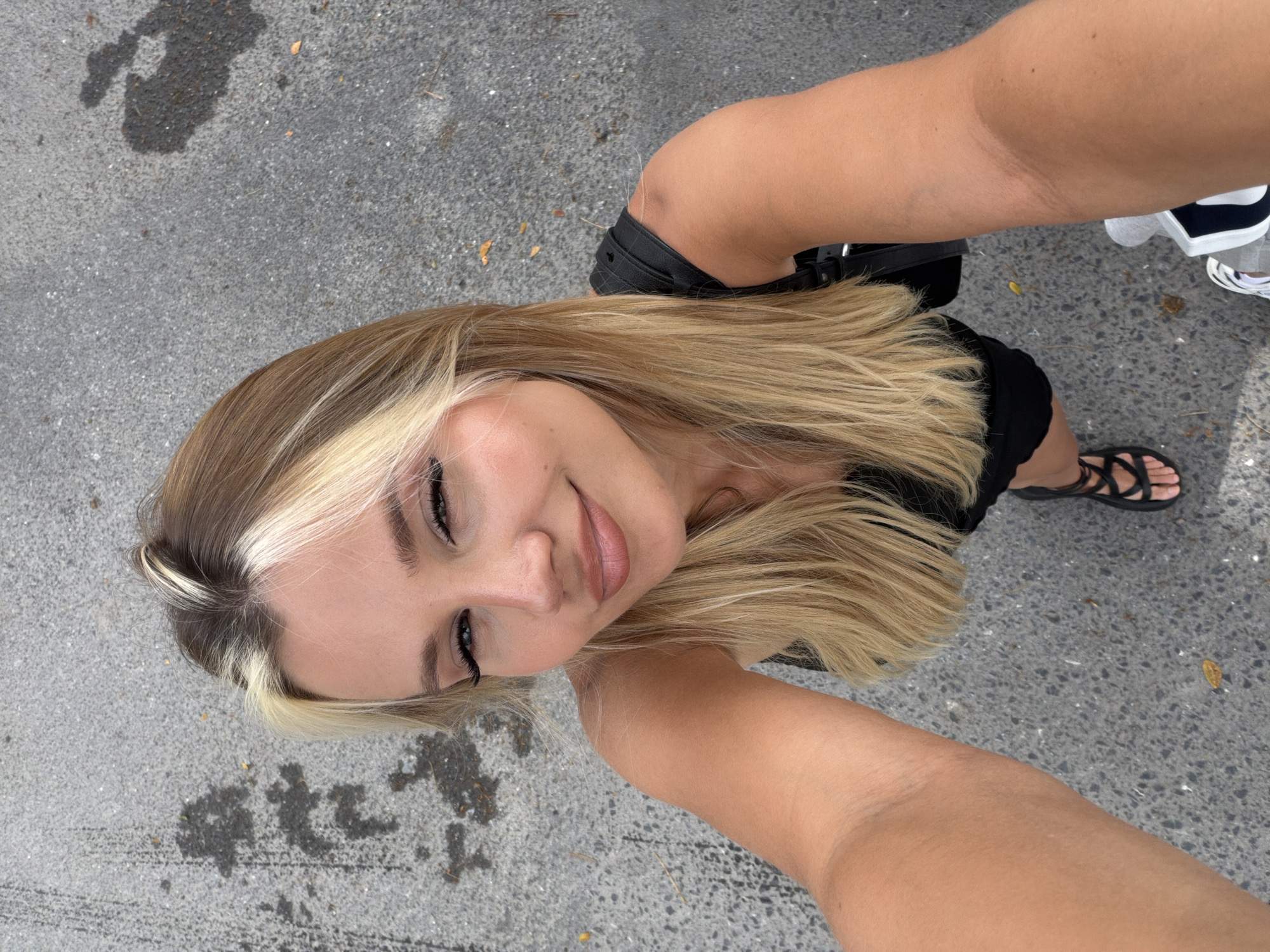 A young woman with blonde hair, styled with blonde streaks, is taking a selfie outdoors. She is wearing a black sleeveless top and black sandals. The ground beneath her appears to be asphalt with some wet patches. The lighting suggests it is daytime. The woman's face is turned upwards towards the camera, and she has a subtle smile. Her eyes are looking directly into the lens, and she is wearing makeup, including eyeliner and mascara. Her hair is long and falls around her shoulders. A black backpack or bag strap is visible over her left shoulder. In the bottom right corner, a person wearing grey athletic shorts and white sneakers with black accents is partially visible, facing away from the camera. The overall setting suggests an urban or semi-urban environment.