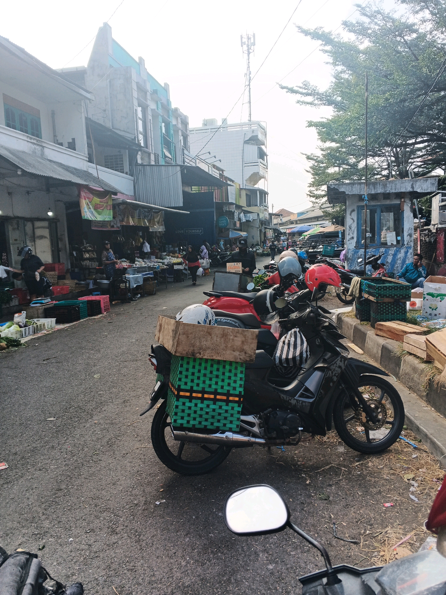 This is a street-level, eye-level photograph of a marketplace scene in Ungaran, Indonesia. The foreground is dominated by a black motorcycle with a large, green woven basket mounted on the rear, filled with what appears to be produce. Two helmets, one red and one striped, are placed on the motorcycle. The motorcycle is parked on an asphalt road.
To the left of the motorcycle, a row of market stalls lines the street. These stalls feature various goods, including crates of vegetables and other merchandise displayed on tables and shelves. Several people are visible in the market area, some attending to their stalls and others browsing.
In the background, several multi-story buildings with varied architectural styles are visible. One building has a distinctive light blue facade at the top. A cell tower is also present in the distance. The overall environment suggests a busy, open-air market with ongoing commercial activity. The lighting indicates it is daytime, with the sky appearing bright and overcast.