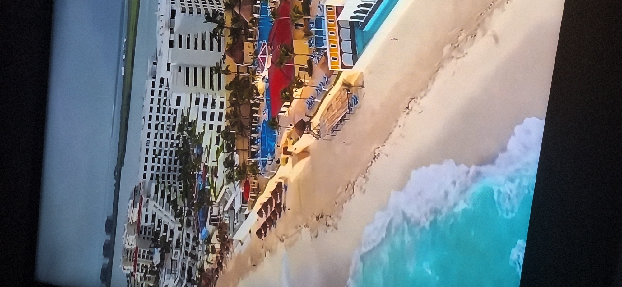 The image is an aerial view of a tropical resort situated along a sandy beach. The resort features multiple white buildings with red-tiled roofs, surrounded by palm trees. There are swimming pools, lounge chairs, and umbrellas visible. The ocean is a vibrant turquoise color with white waves crashing onto the shore. In the distance, across a body of water, other structures and landmasses are visible.
This image does not depict the city of Bila Tserkva, Ukraine. Bila Tserkva is an inland city located in central Ukraine, far from any tropical coastal resorts and oceans.