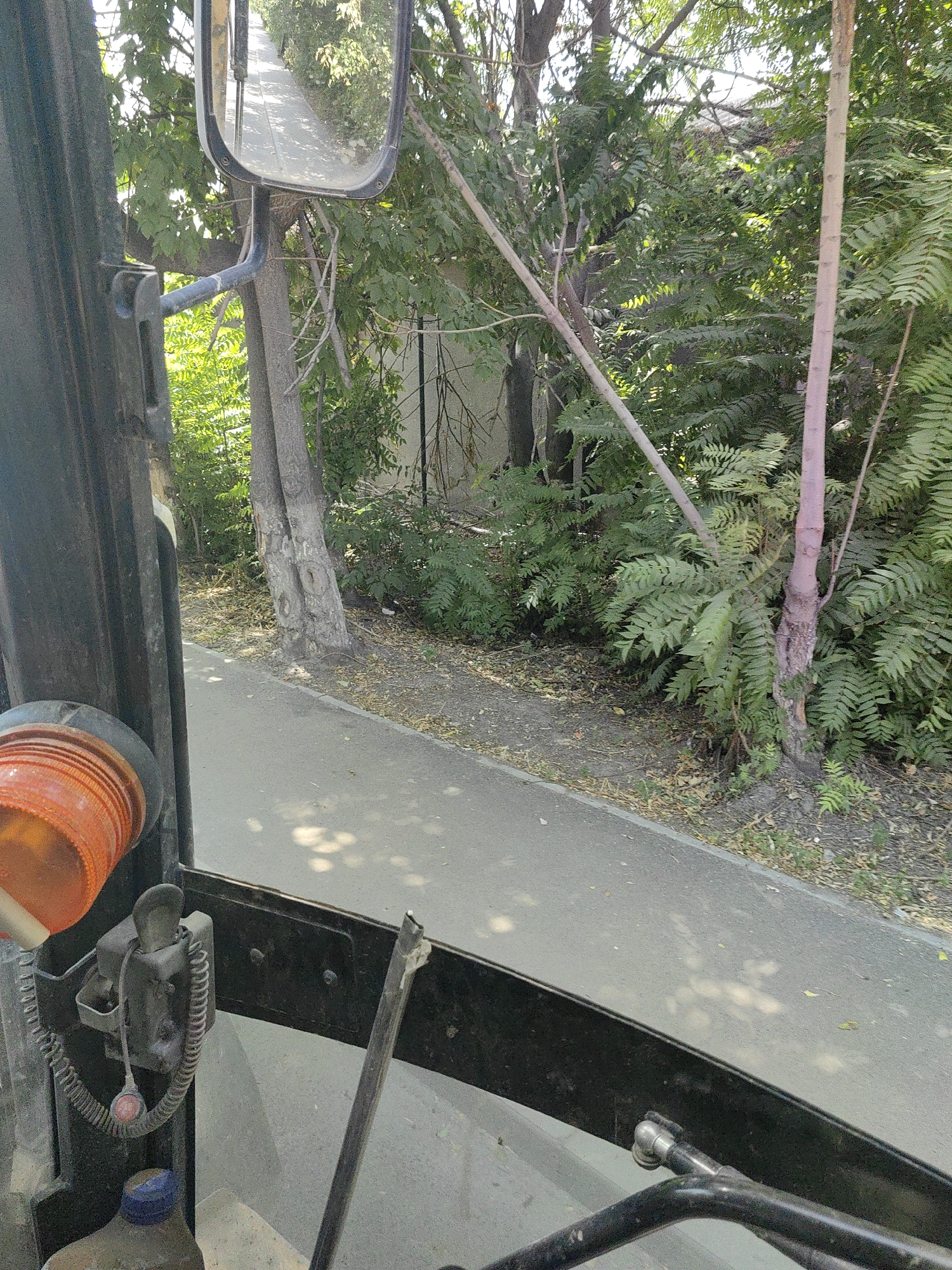 The image captures a view from inside a vehicle, likely a forklift or similar machinery, looking out through the windshield. In the foreground, a portion of the vehicle's interior is visible, including an orange warning light and some control mechanisms. Outside, a paved pathway runs alongside lush green vegetation and trees. A side mirror is mounted on the vehicle, reflecting a road or path receding into the distance, bordered by more trees. The scene appears to be in an outdoor urban or semi-urban setting, possibly a park or a street with abundant greenery. The lighting suggests it is daytime, with sunlight filtering through the leaves, creating dappled shadows on the pathway. The context of Shymkent, Kazakhstan, is not visually discernible from the image itself; it is external information provided as a location.