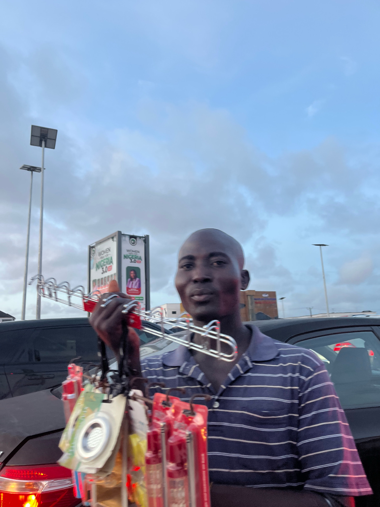 A man is standing in a car park, holding a rack of small items, possibly for sale. The man is bald and wearing a blue and white striped polo shirt. He is looking towards the camera with a slight smile. In the background, there are cars, streetlights, and a billboard advertising "Women Nigeria 3.0". The sky is cloudy and overcast, indicating it might be evening or a cloudy day. The location is identified as Lekki Peninsula II, Nigeria.