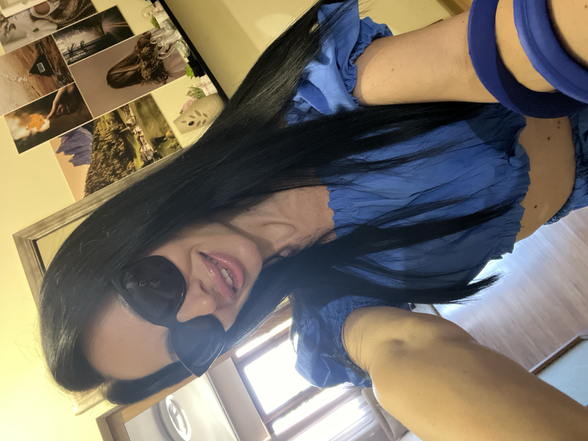 A medium shot shows a woman from the chest up, posing for a selfie. She has long, black hair and is wearing large, dark sunglasses and a blue off-the-shoulder top with puffy sleeves. Her lips are glossed and slightly parted as she smiles. Her left arm is bent, and a dark blue bangle is visible on her wrist. The background is a room with a yellow wall and a shelf displaying framed pictures of landscapes and a woman. A window with wooden blinds is visible behind her to the left. The flooring appears to be wood. The image quality is good, with natural lighting. The woman's pose suggests she is indoors, likely in a residential setting. There is no indication of the specific location within the city of Stara Zagora, Bulgaria.