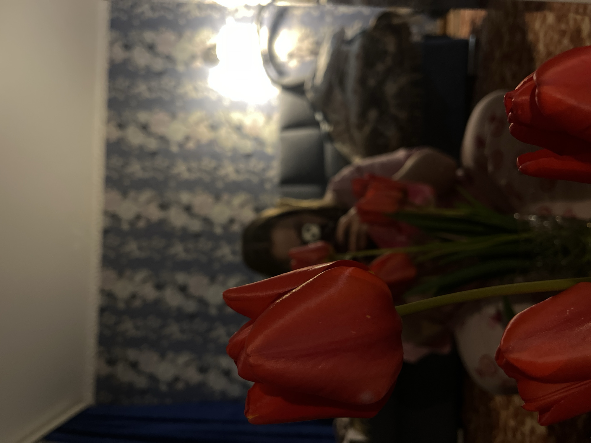 The image, taken in Novopokrovskaya, Russia, depicts an indoor scene with a shallow depth of field, focusing sharply on several vibrant red tulips in the foreground and mid-ground. The tulips, with their rich red petals and green stems, are arranged, possibly in a vase which is largely obscured.
In the blurred background, a person, appearing to be a young individual or adult with long dark hair, is partially visible. They are wearing a light-colored top and seem to be looking towards or interacting with the flowers. Behind the person, a dark piece of furniture, possibly a sofa or bed, can be discerned. The wall in the background features patterned wallpaper with blue, grey, and lighter, abstract or cloud-like designs.
A very bright, out-of-focus light source, likely an indoor lamp, is prominent in the upper left of the image, creating a strong glow and soft bokeh effects. The lighting suggests an interior setting, possibly in the evening or at night, where artificial light is dominant. The overall atmosphere is domestic and quiet, capturing a moment of appreciation or arrangement of the flowers. There is no visible text in the image.