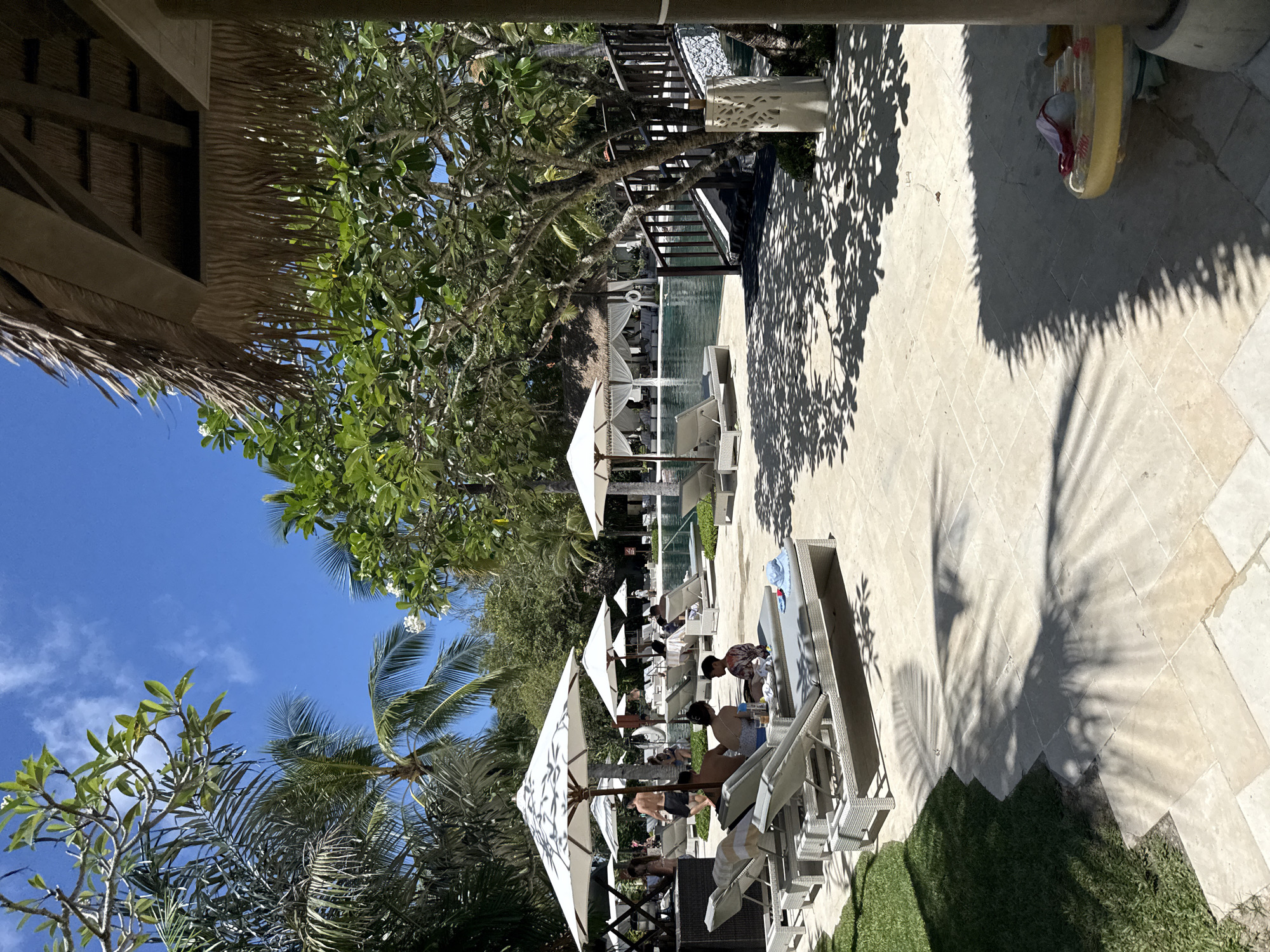 The image captures a poolside scene in Nusa Dua, Indonesia. People are relaxing on lounge chairs under white umbrellas, positioned along a pool. Trees and palm trees provide shade and add to the tropical ambiance. The sky is clear and blue. A building with a thatched roof is partially visible on the left. The foreground is a paved area.