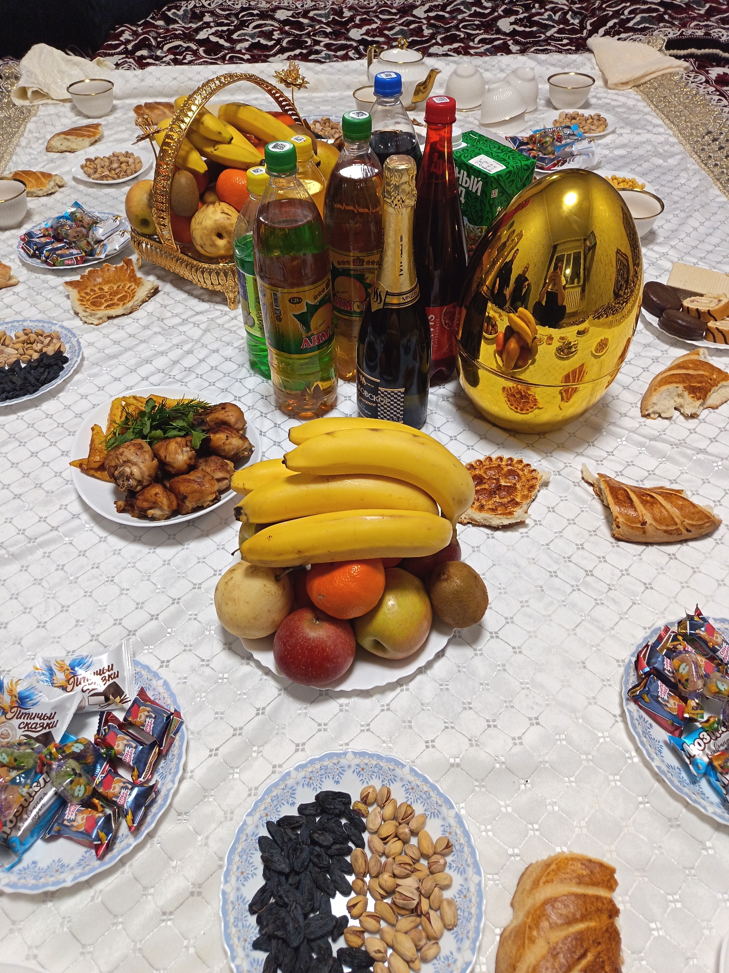 A high-angle shot captures a table laden with food and drinks. The table is covered with a white, patterned tablecloth. Various dishes and items are arranged on the table.
A golden basket filled with bananas, oranges, kiwis, and pears is partially visible. There are also plates of bread, nuts, and candies. A golden, egg-shaped object reflects the surroundings, including what appears to be a person taking the photo.
Bottles of drinks are interspersed among the food items, including bottles labeled in Cyrillic characters. Plates of fried chicken and potatoes are arranged towards the left side of the table.
The setting is indoors, likely a home or a banquet hall, based on the presence of the tablecloth and food arrangement. Given the Cyrillic labeling on the bottles, it is likely in a country where that alphabet is used, potentially Russia. There is no information to determine if this image was taken in Nizhnevartovsk, Russia.